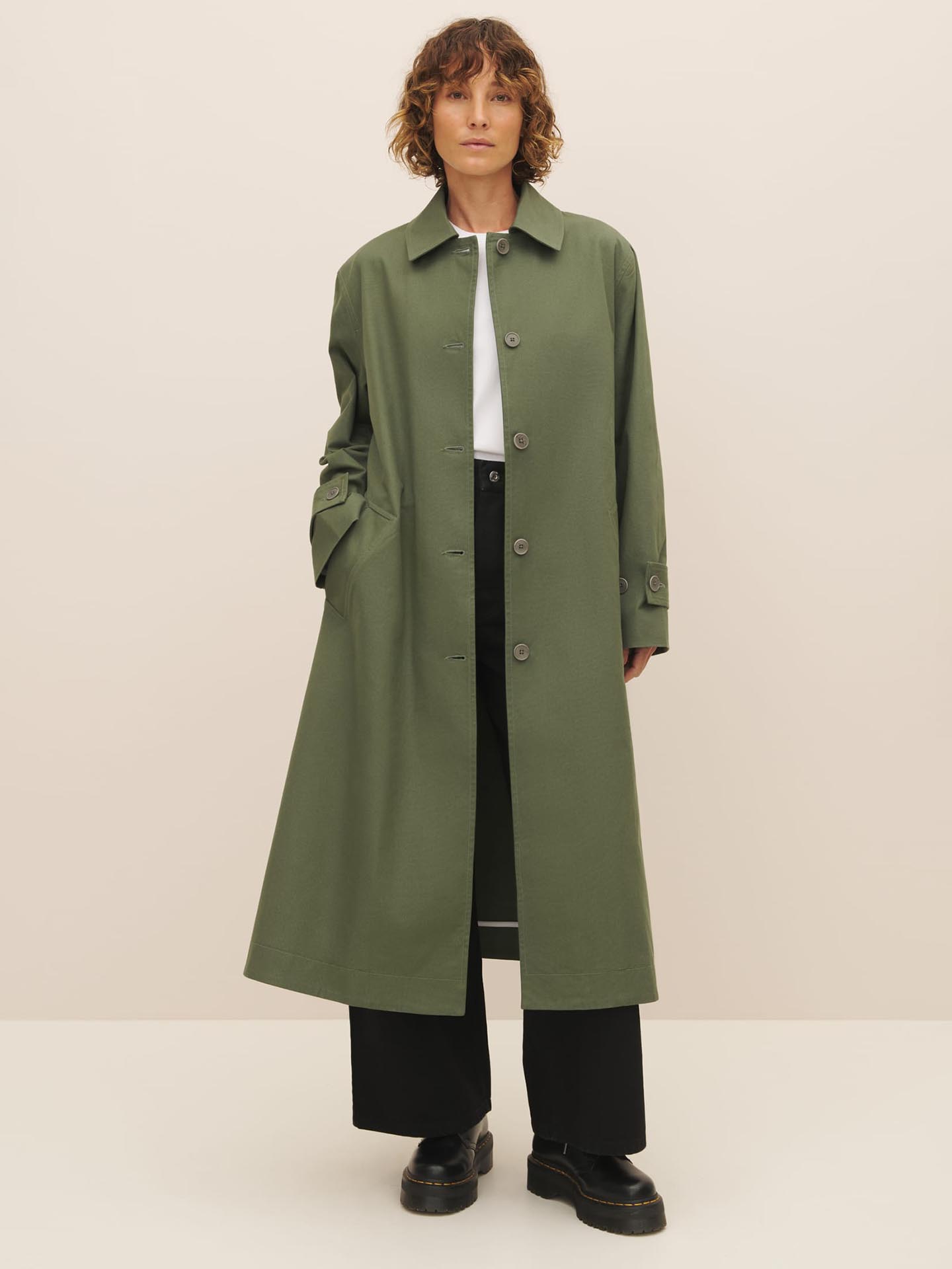 Sentence with replacement: A person wearing a Kowtow Cleo Trench Coat in Sage, black pants, and black shoes against a neutral background.