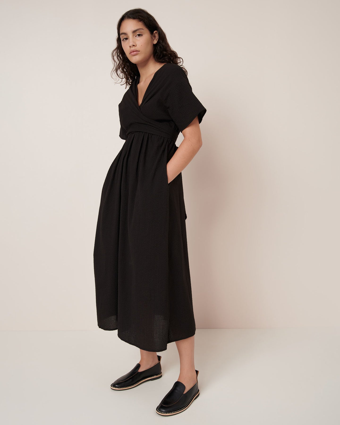A woman stands against a plain background wearing the Kowtow Composer Dress – Black, a short-sleeved, v-neck, ankle-length dress with pockets and black slip-on shoes. Standard fit—see our size guide for detailed measurements.