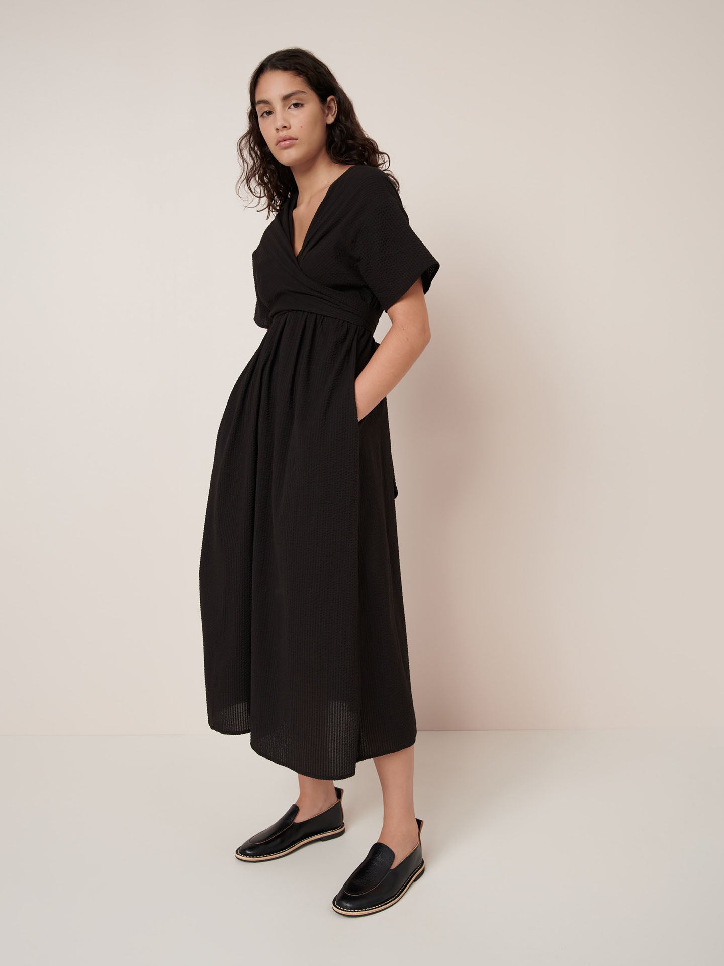 A woman stands against a plain background wearing the Kowtow Composer Dress – Black, a short-sleeved, v-neck, ankle-length dress with pockets and black slip-on shoes. Standard fit—see our size guide for detailed measurements.