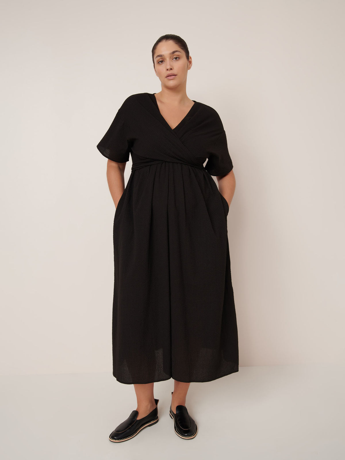 A woman stands against a plain background wearing the Kowtow Composer Dress – Black, a short-sleeve, V-neck, ankle-length dress with a standard fit, paired with black slip-on shoes. Consult our sizing guide for your perfect fit.