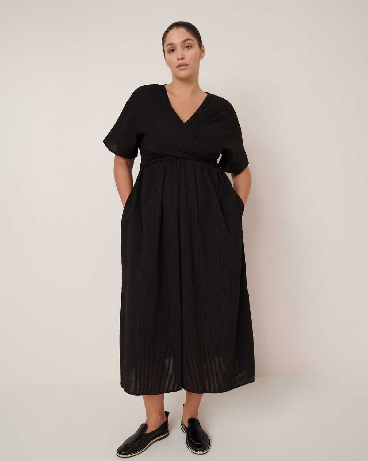 A woman stands against a plain background wearing the Kowtow Composer Dress – Black, a short-sleeve, V-neck, ankle-length dress with a standard fit, paired with black slip-on shoes. Consult our sizing guide for your perfect fit.