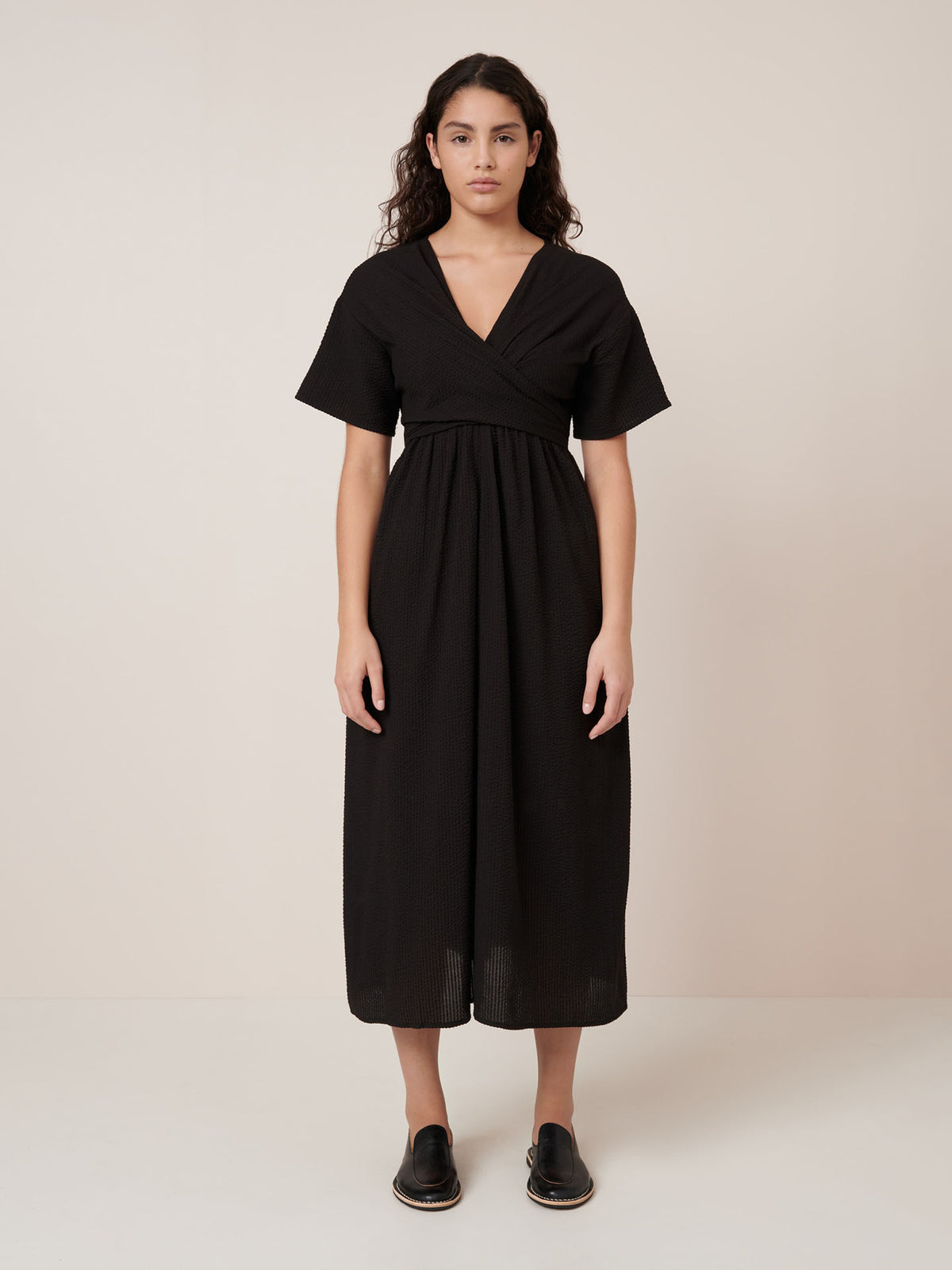 A woman stands against a plain light background wearing Kowtow&#39;s Composer Dress in black—an ankle-length, short-sleeve style with a standard fit—paired with black slip-on shoes. See our size guide for garment measurements.