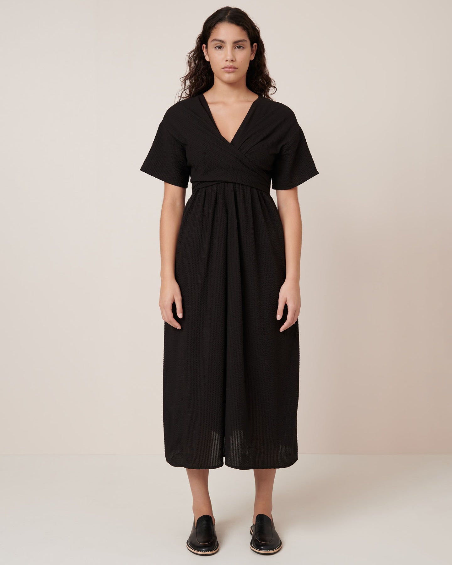 A woman stands against a plain background wearing the Kowtow Composer Dress – Black, a short-sleeved, v-neck, ankle-length dress with pockets and black slip-on shoes. Standard fit—see our size guide for detailed measurements.