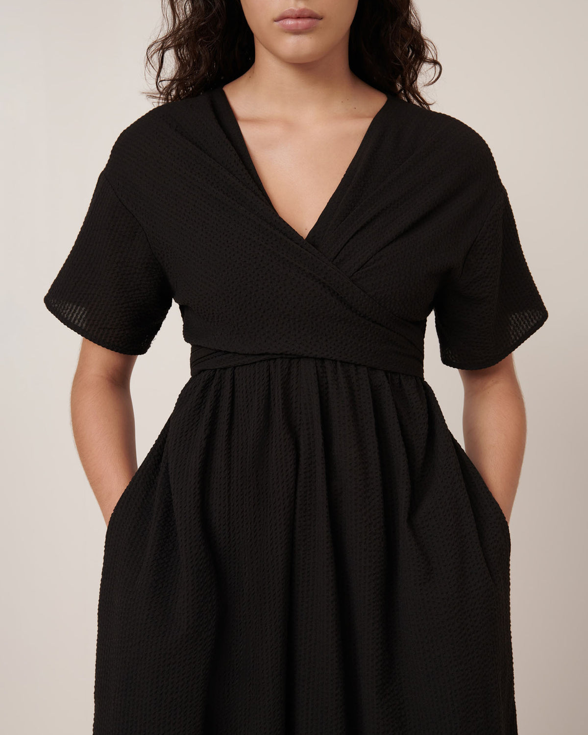 A woman wears the Kowtow Composer Dress – Black, a short-sleeved, textured wrap style with V-neckline and pockets. Check our size guide for garment measurements to find your perfect fit.