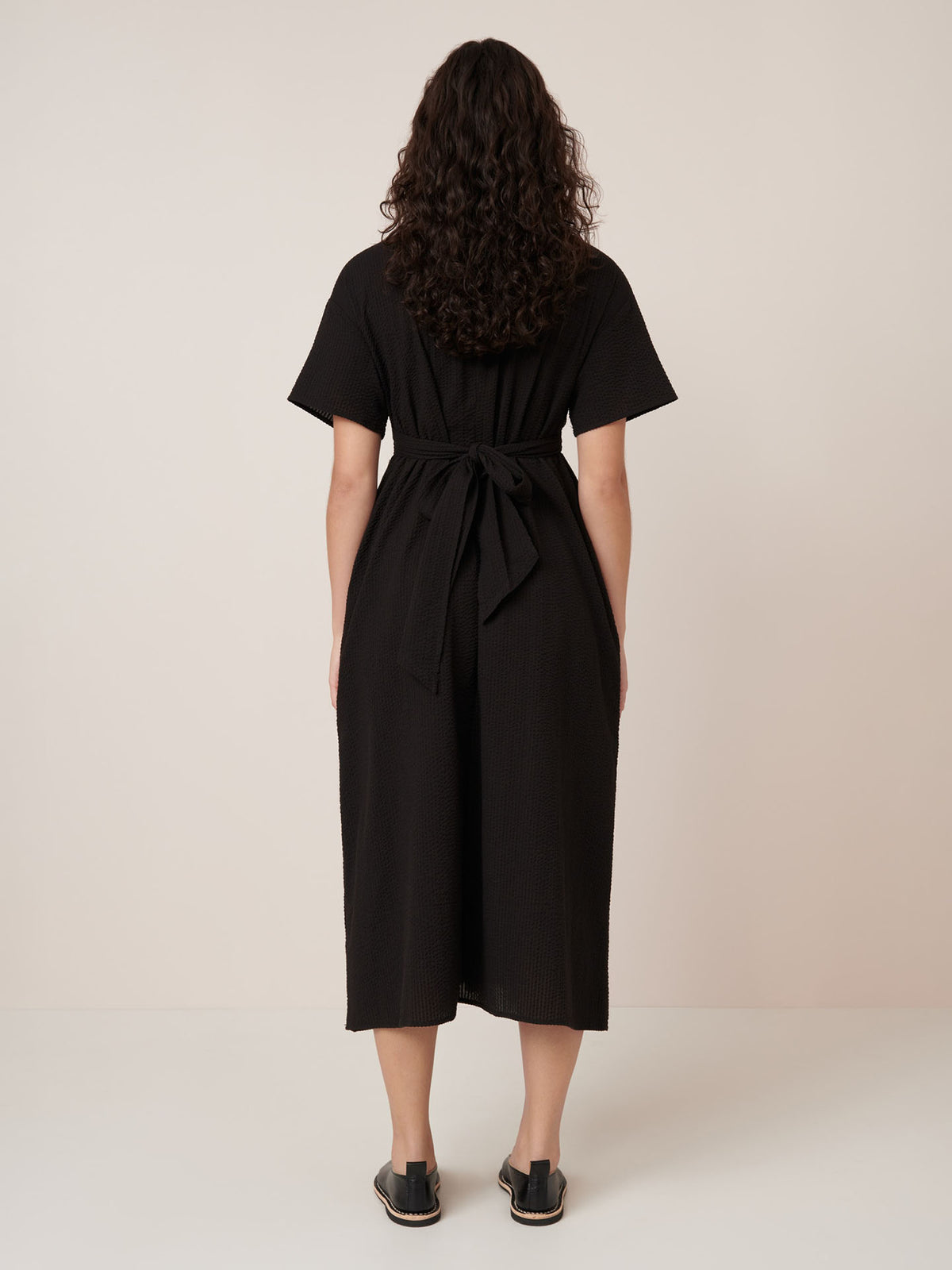 A woman with curly hair stands facing away, wearing the Kowtow Composer Dress – Black, a belted, short-sleeve midi dress in a standard fit, paired with black slip-on sandals against a plain light background.