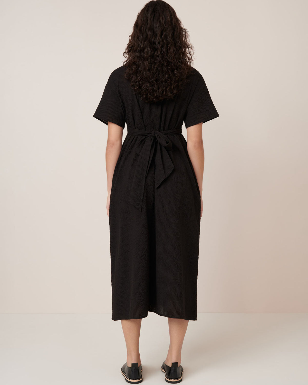 A woman with curly hair stands facing away, wearing the Kowtow Composer Dress – Black, a belted, short-sleeve midi dress in a standard fit, paired with black slip-on sandals against a plain light background.