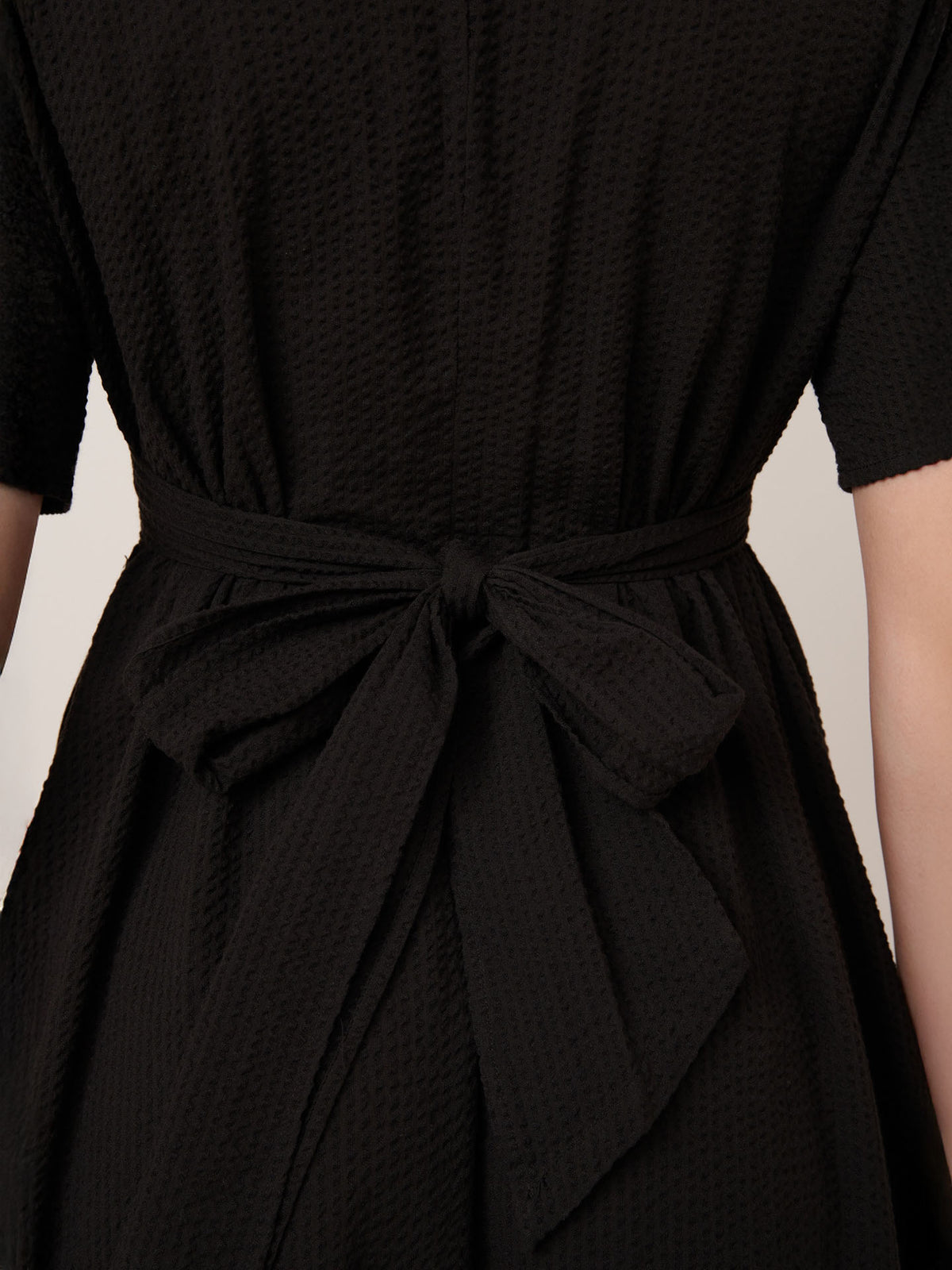 Close-up of the back of a person wearing Kowtow’s Composer Dress in Black, featuring a textured fabric and matching belt tied at the waist in their standard fit.
