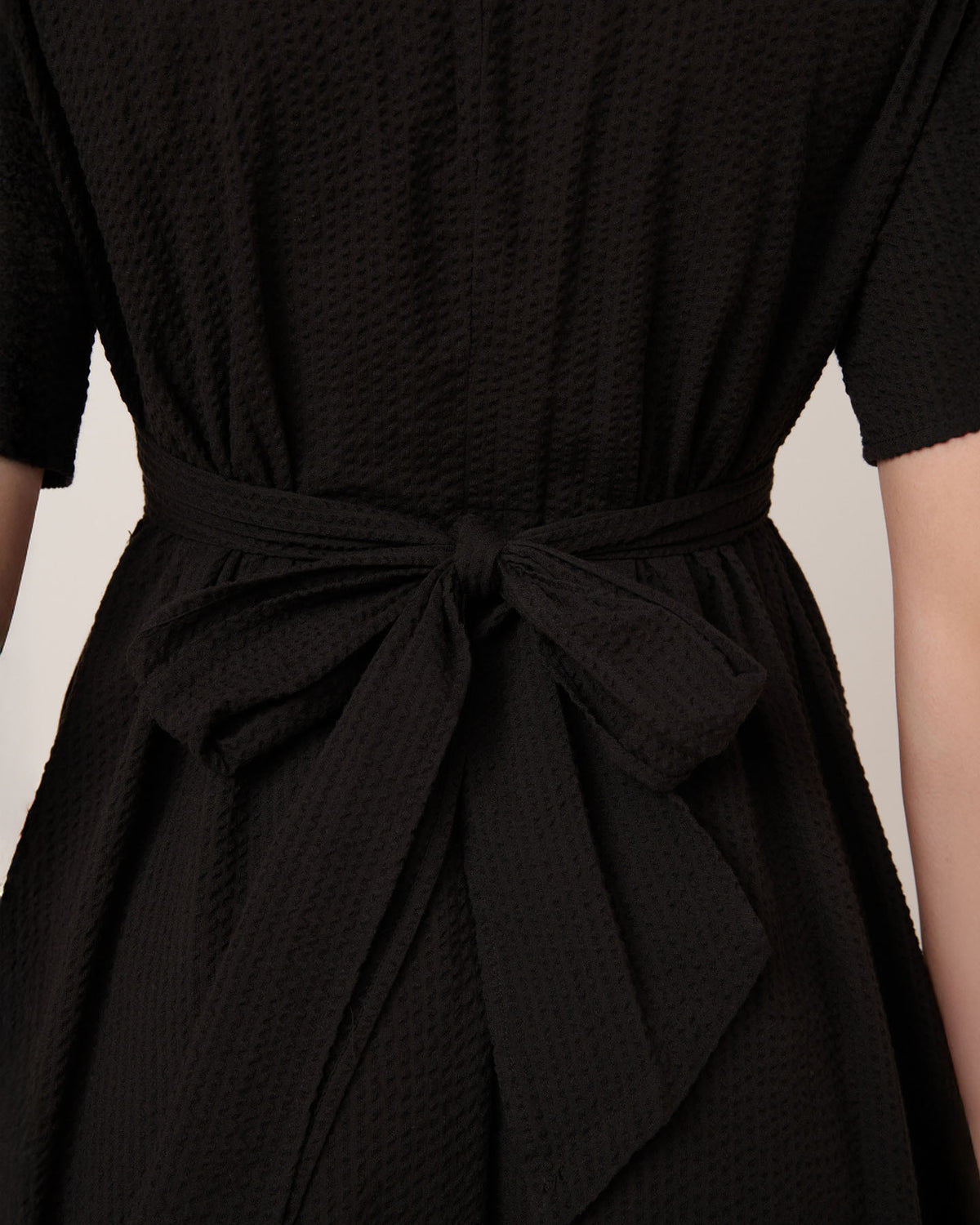 Close-up of the back of a person wearing Kowtow’s Composer Dress in Black, featuring a textured fabric and matching belt tied at the waist in their standard fit.