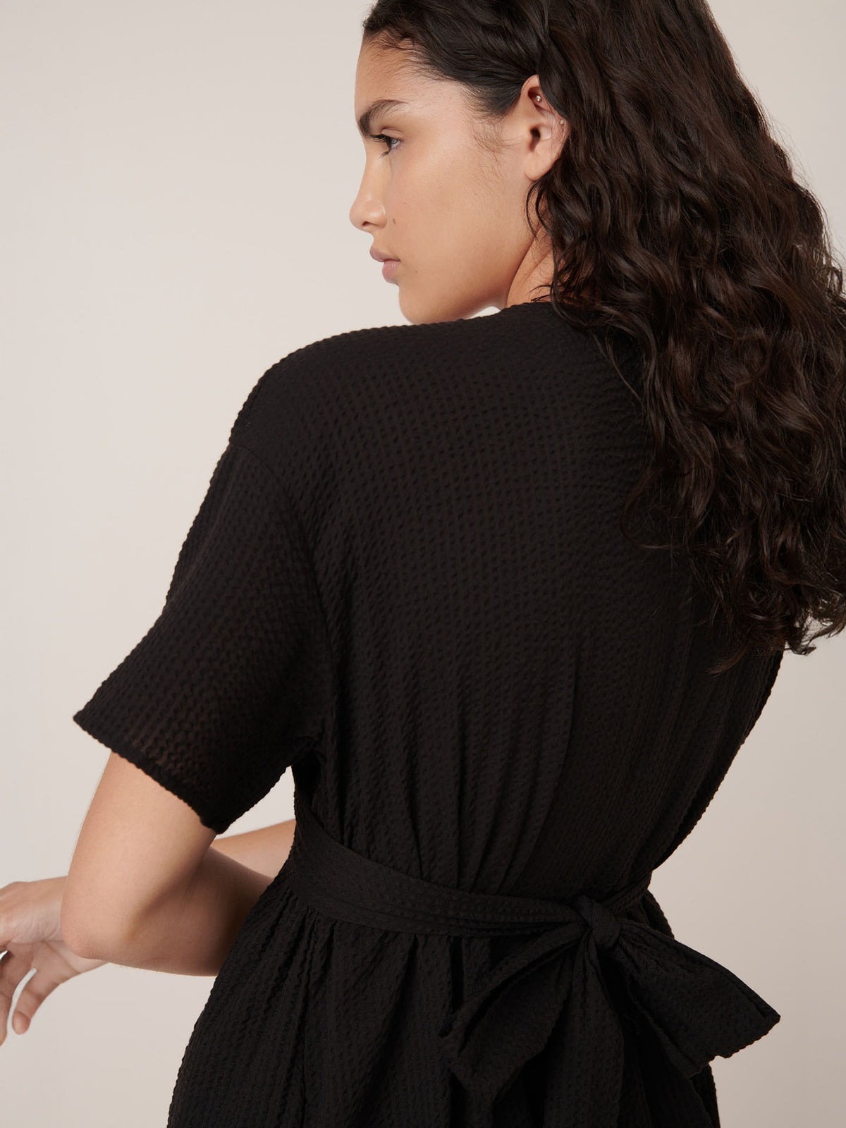 A woman with wavy dark hair faces away, looking left, wearing the Kowtow Composer Dress in black—a textured style with a standard fit and waist tie.