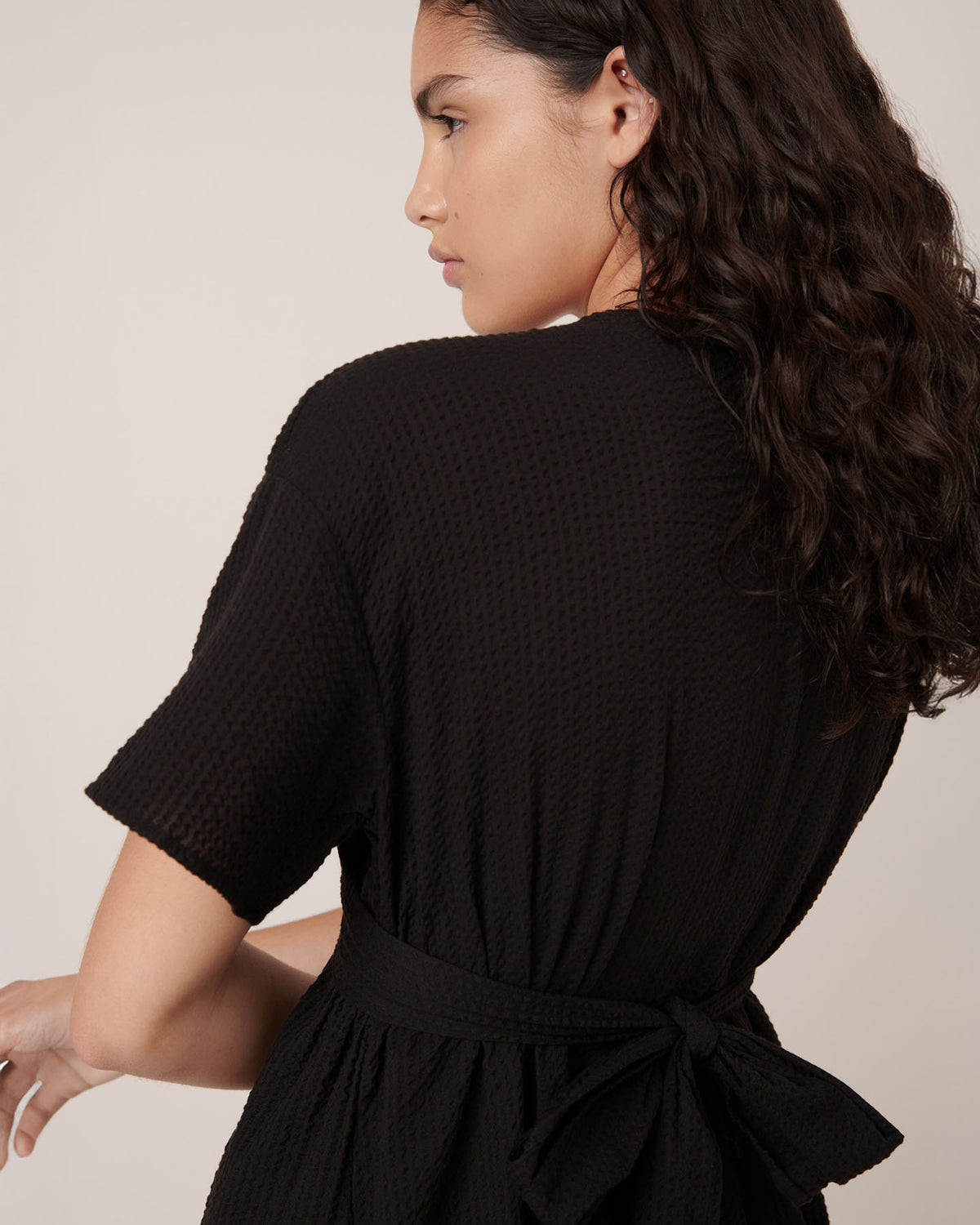 A woman with wavy dark hair faces away, looking left, wearing the Kowtow Composer Dress in black—a textured style with a standard fit and waist tie.