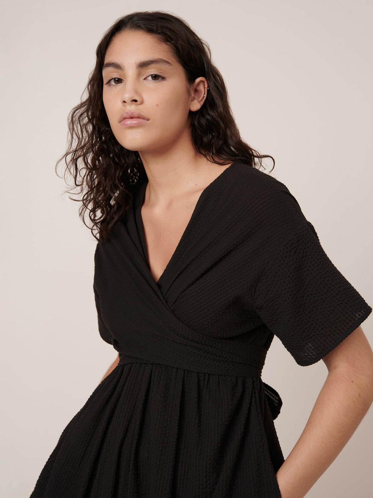 A person with wavy dark hair wears a short-sleeved black Kowtow Composer Dress in a standard fit, standing against a plain light background and looking at the camera.