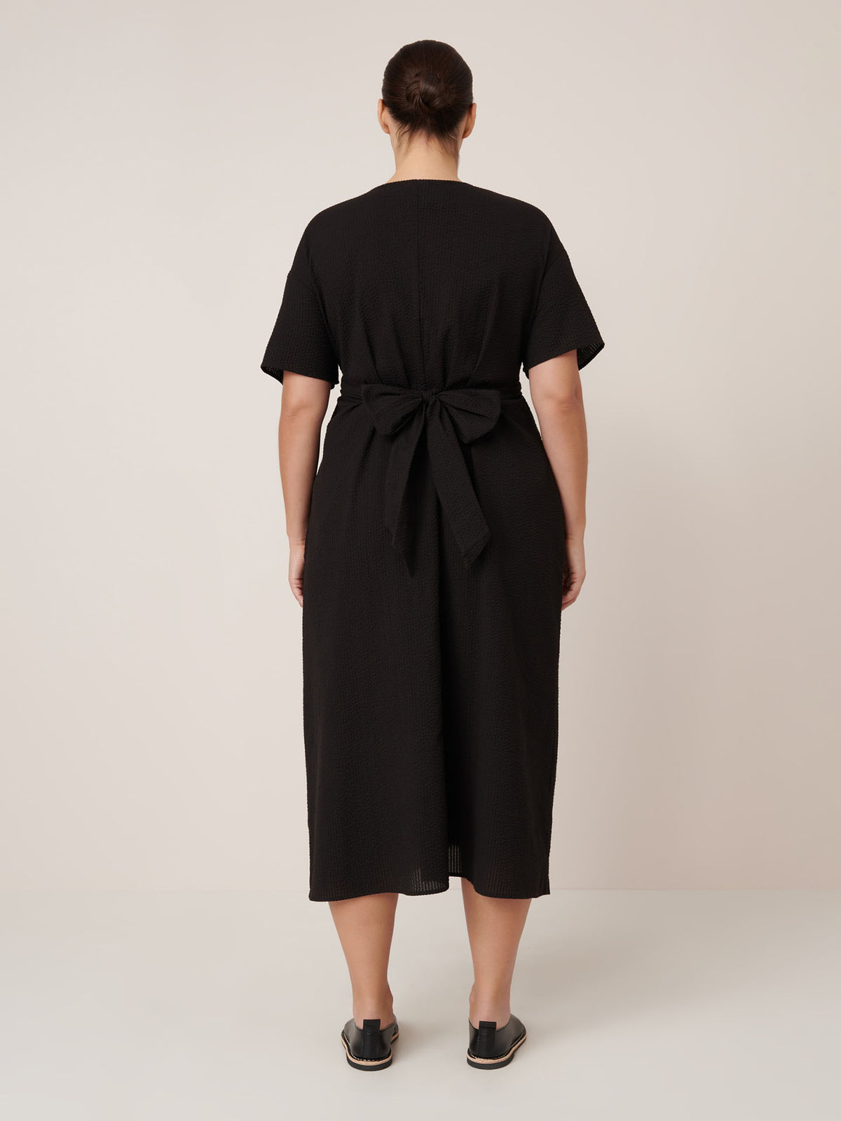 A woman faces away, wearing the Kowtow Composer Dress in black—a short-sleeve, waist-tie midi dress with a standard fit—paired with black slip-on shoes against a plain light backdrop. Check our sizing guide for the perfect fit.