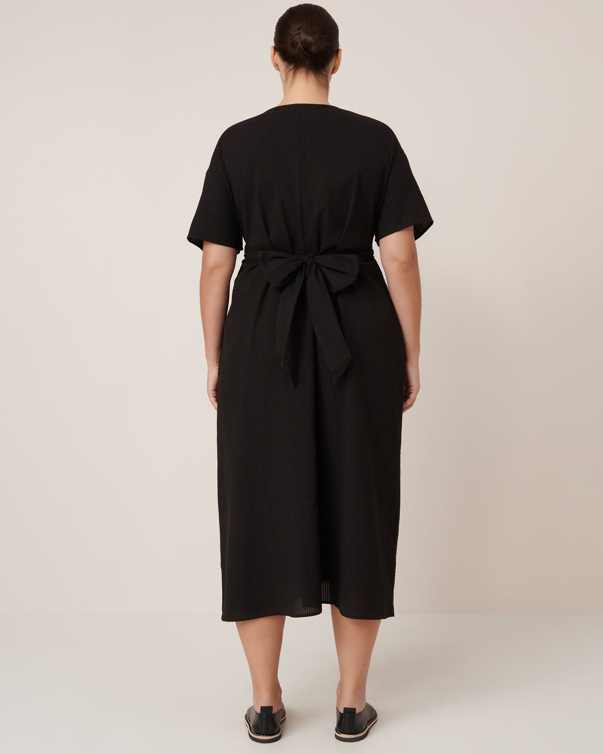A woman faces away, wearing the Kowtow Composer Dress in black—a short-sleeve, waist-tie midi dress with a standard fit—paired with black slip-on shoes against a plain light backdrop. Check our sizing guide for the perfect fit.