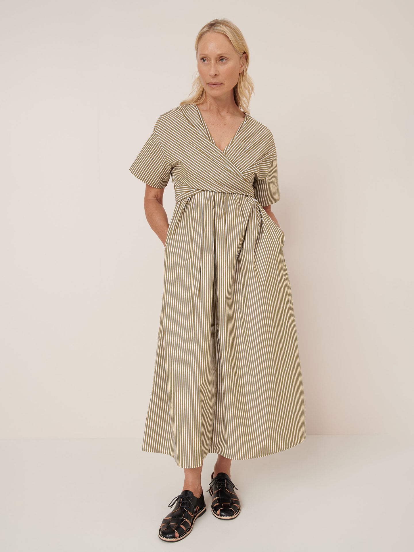 A woman in Kowtow's Composer Dress – Treetop Stripe, a standard fit, short-sleeved, ankle-length beige wrap dress with vertical stripes, stands against a plain background with one hand in her pocket wearing black sandals. See our size guide.