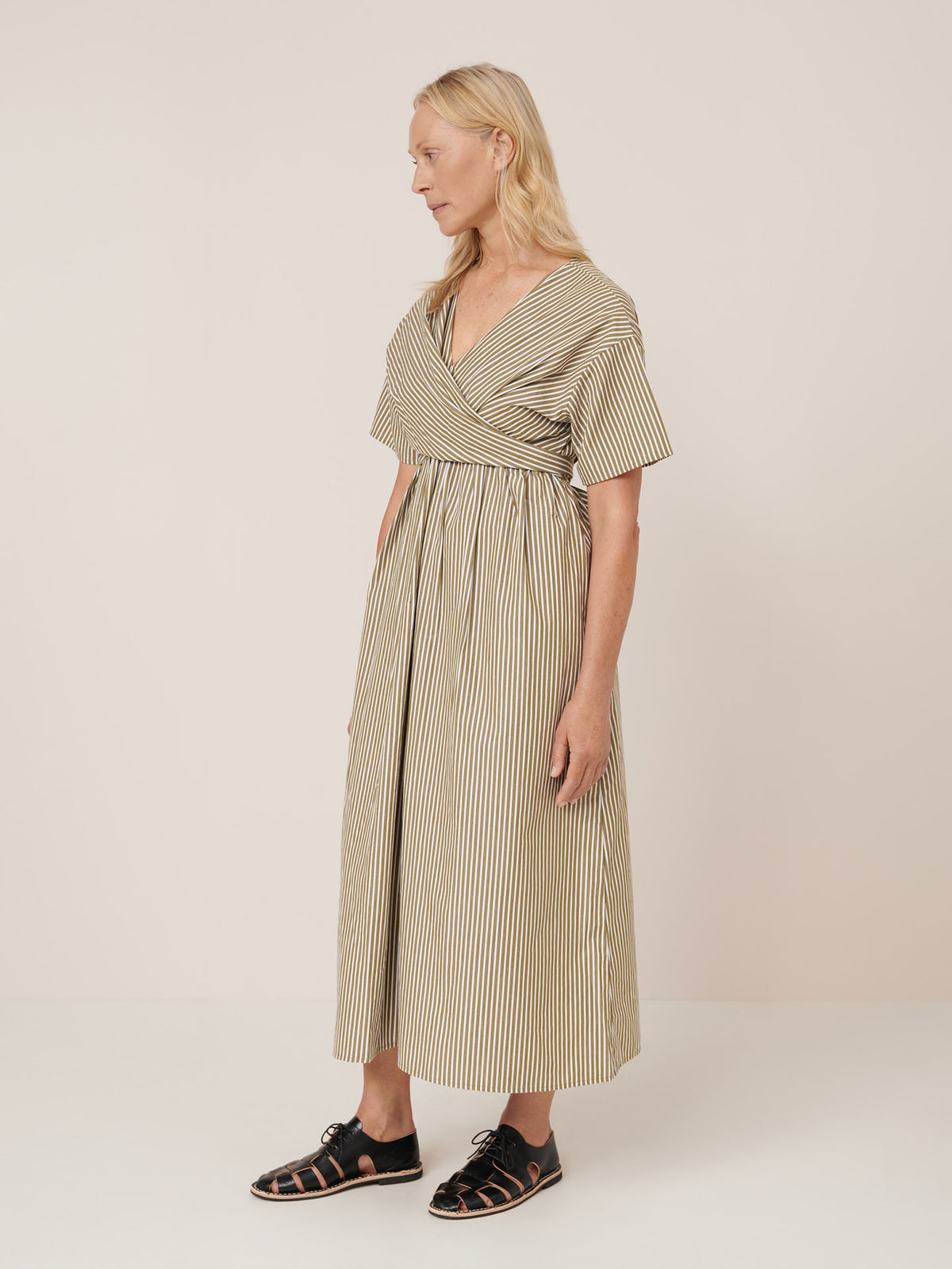 A woman stands in profile wearing the Kowtow Composer Dress – Treetop Stripe, a long, short-sleeved, striped wrap dress, with black sandals against a plain light background. Check our size guide for standard fit and measurements.