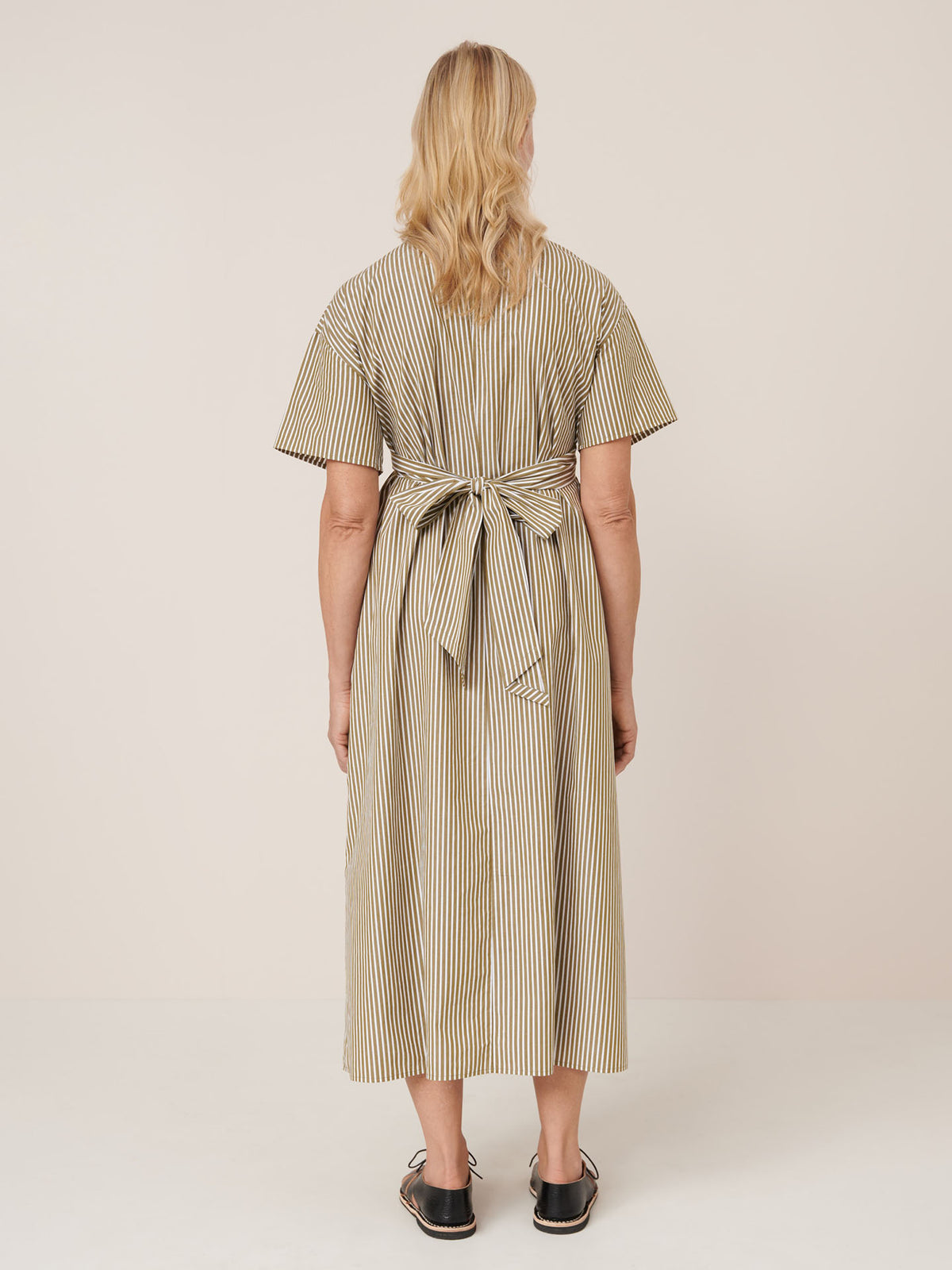 A woman wears the Kowtow Composer Dress – Treetop Stripe: a short-sleeved, ankle-length striped dress with a large back bow, styled with black slip-ons. Check our size guide to find your perfect standard fit.