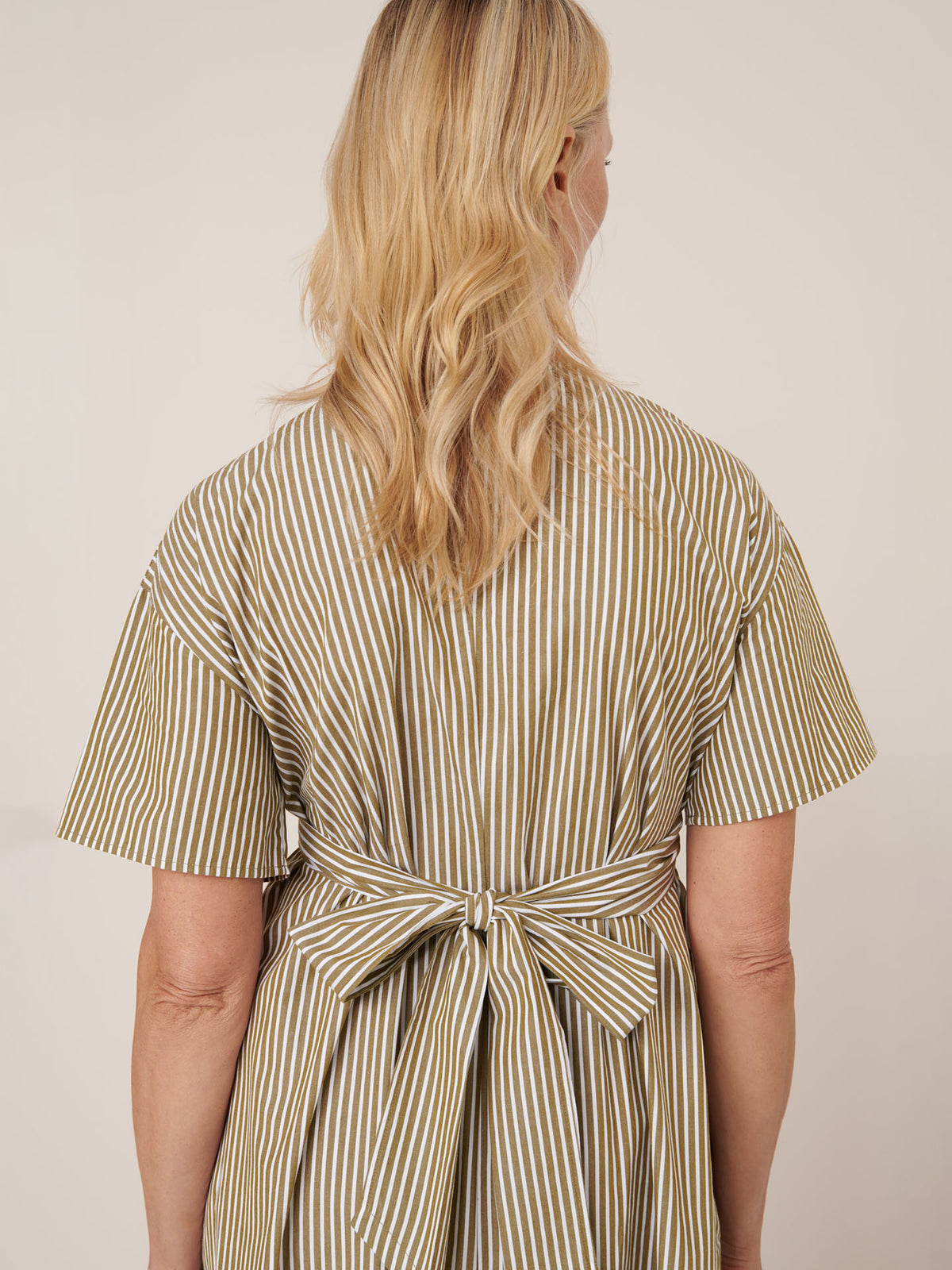 A person with blonde wavy hair faces away, wearing the Kowtow Composer Dress – Treetop Stripe, a standard fit, short-sleeved striped dress with a tied bow at the back. Please check the size guide for garment measurements.