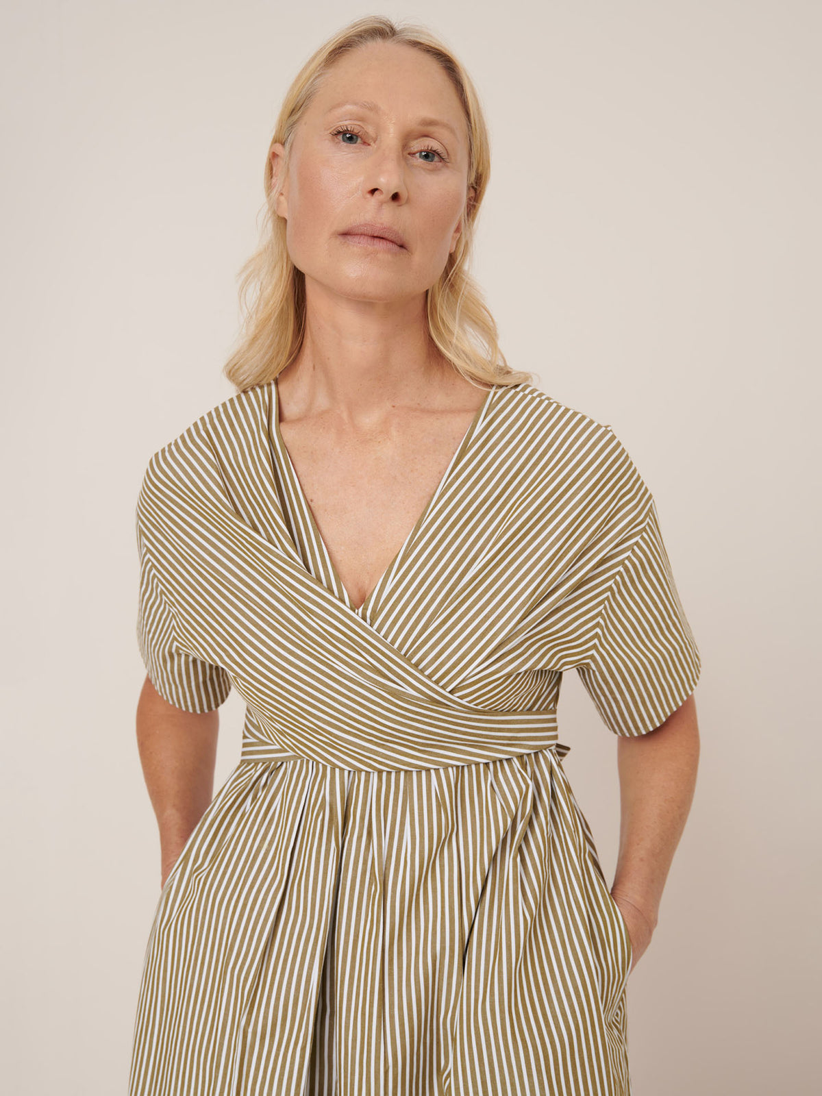 A woman with blonde hair stands with her hands in her pockets, wearing the Kowtow Composer Dress – Treetop Stripe, a standard fit, short-sleeved, V-neck striped dress, against a plain background.