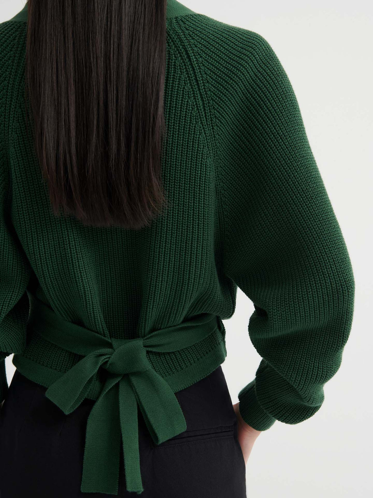 A person wearing the Composure Cardigan – Forest, a dark green knit sweater with a tied bow at the back, paired with black pants. Only the back view is visible. See our sizing guide for your perfect fit.