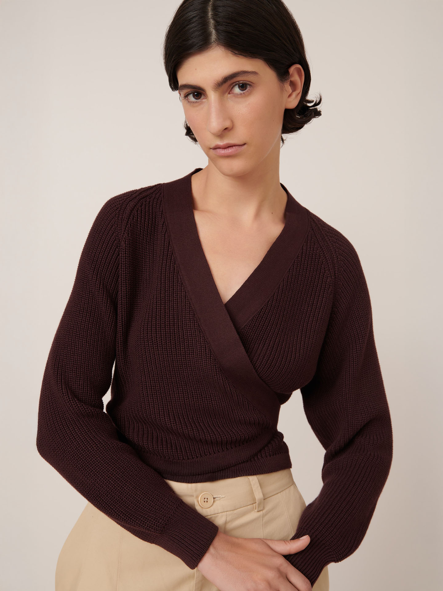 A person with short dark hair wears the Kowtow Composure Cardigan in Mahogany with beige high-waisted pants, standing against a plain light background. The standard fit highlights clean lines and effortless style.
