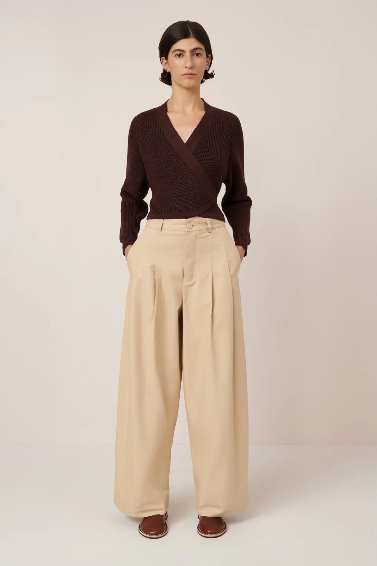A person stands against a plain background wearing Kowtow’s Composure Cardigan in Mahogany, paired with wide-leg beige trousers and brown slip-on shoes, hands in pockets. The standard fit offers comfort without sacrificing style.