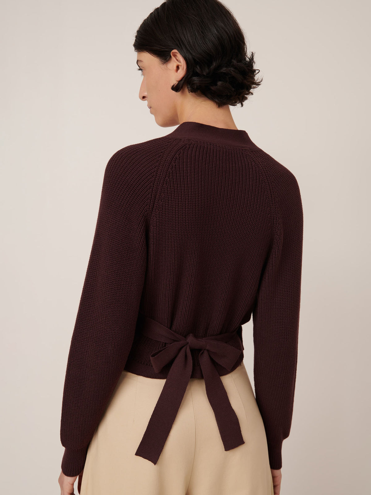 A woman is seen from behind wearing the Kowtow Composure Cardigan in mahogany, featuring a back bow tie and standard fit, paired with beige pants against a plain background.