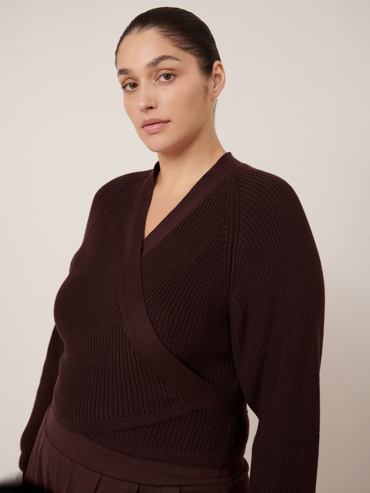 A person with dark hair pulled back is wearing the Kowtow Composure Cardigan in Mahogany, a long-sleeve knitwear wrap, along with matching pants, standing against a plain light background.