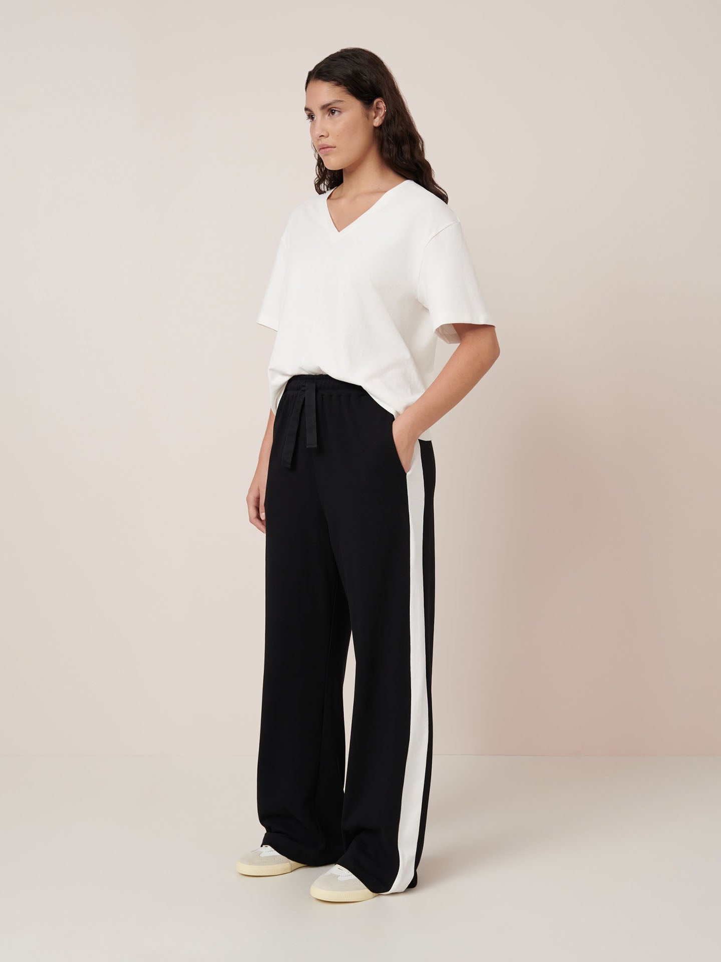 A person stands against a plain background wearing a Kowtow white V-neck short-sleeve top, Kowtow Contrast Trackpant – Black with white side stripes, and white shoes.