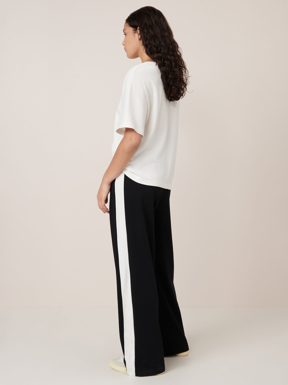 A person stands sideways wearing a relaxed fit white Kowtow T-shirt and Kowtow’s Contrast Trackpant in black, featuring white side stripes, against a plain light background.