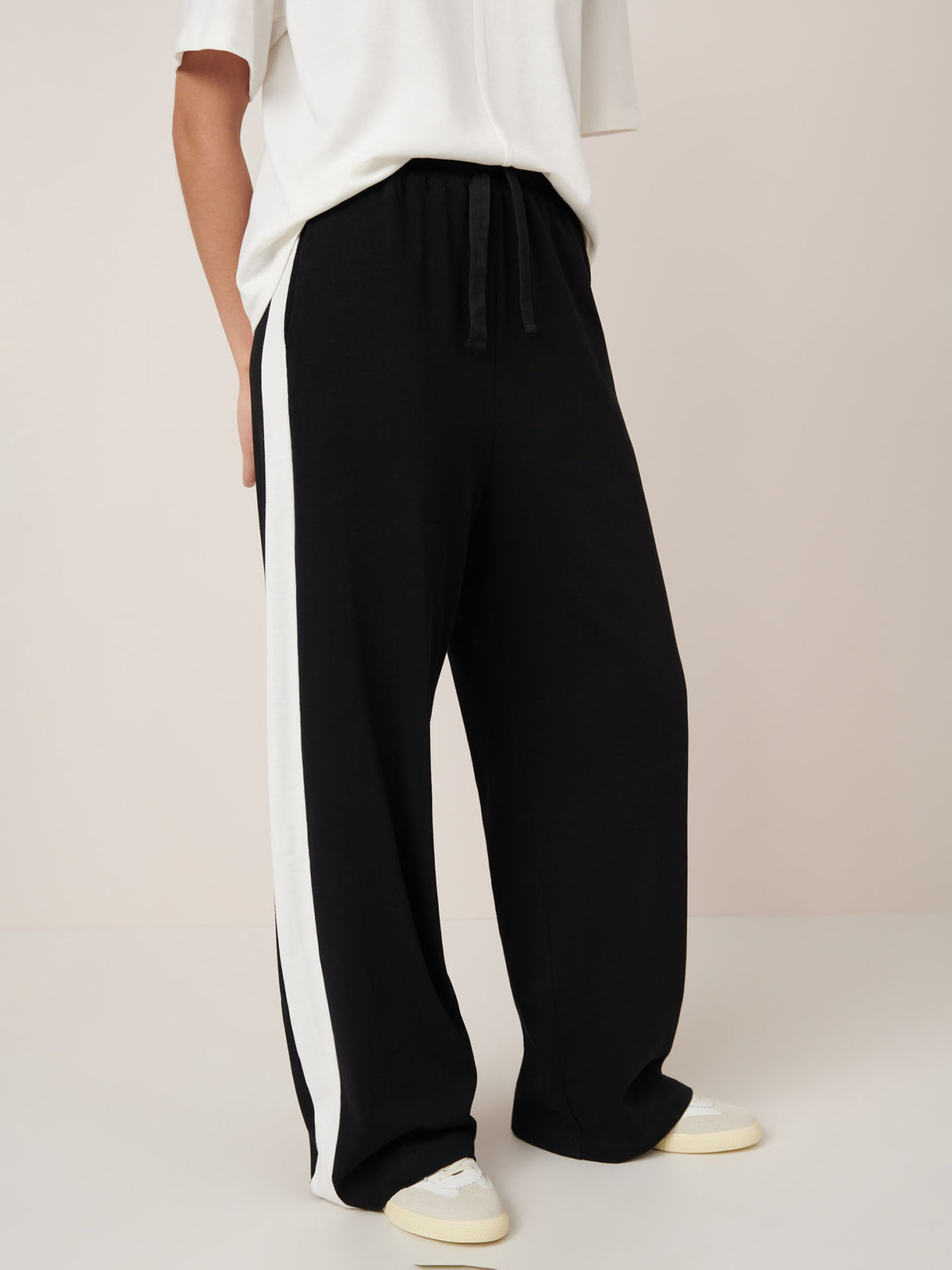 Model wears the Kowtow Contrast Trackpant – Black, a relaxed fit, high waisted pant with a white side stripe, paired with a white top and sneakers against a plain background.