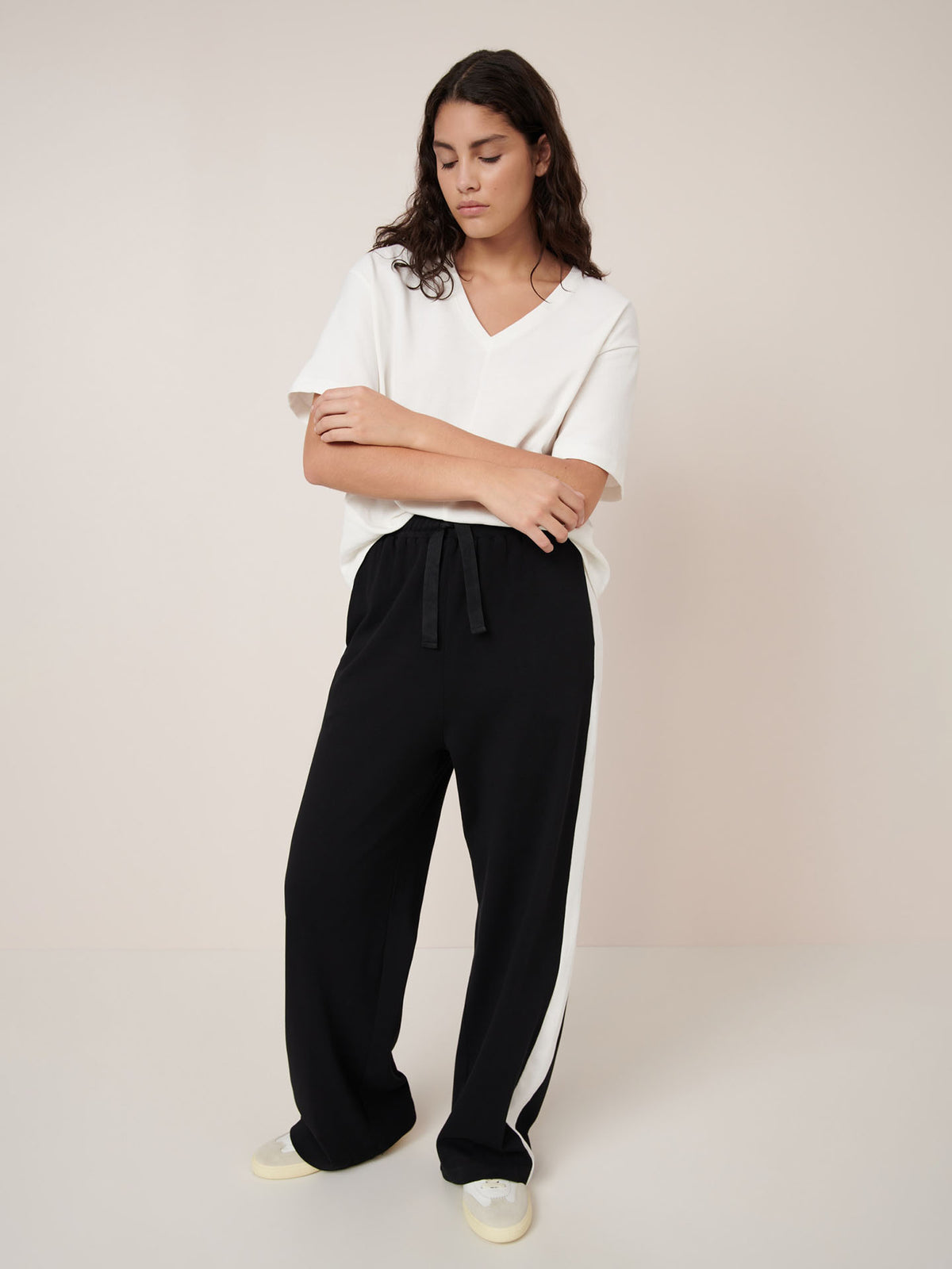 A person stands against a plain background wearing a relaxed fit white V-neck top, Kowtow Contrast Trackpant – Black with white side stripes, and light-colored shoes.