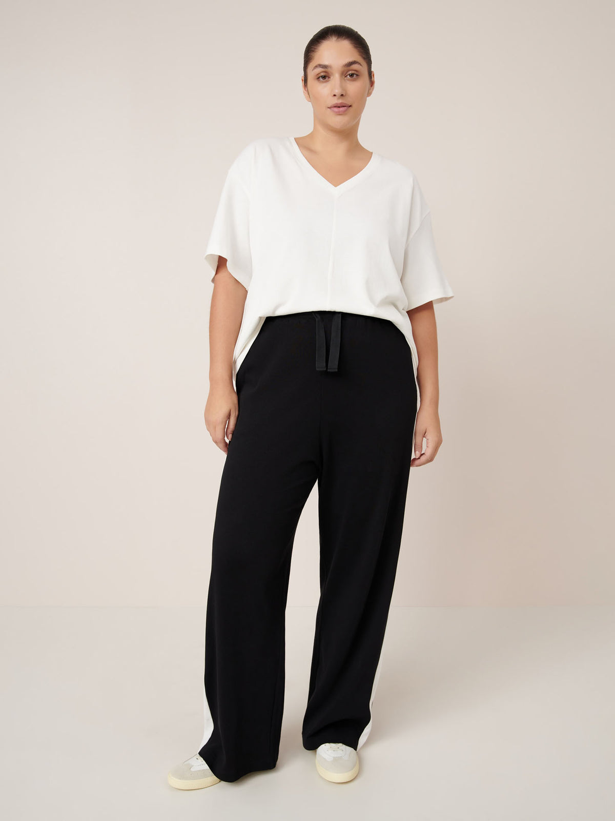 A person stands against a plain background wearing a white V-neck top, Kowtow Contrast Trackpant – Black in a relaxed fit with a drawstring, and white sneakers.