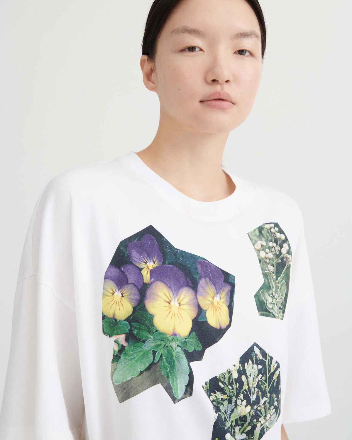 A white t-shirt featuring graphic cutouts of purple and yellow pansies and green wildflowers.