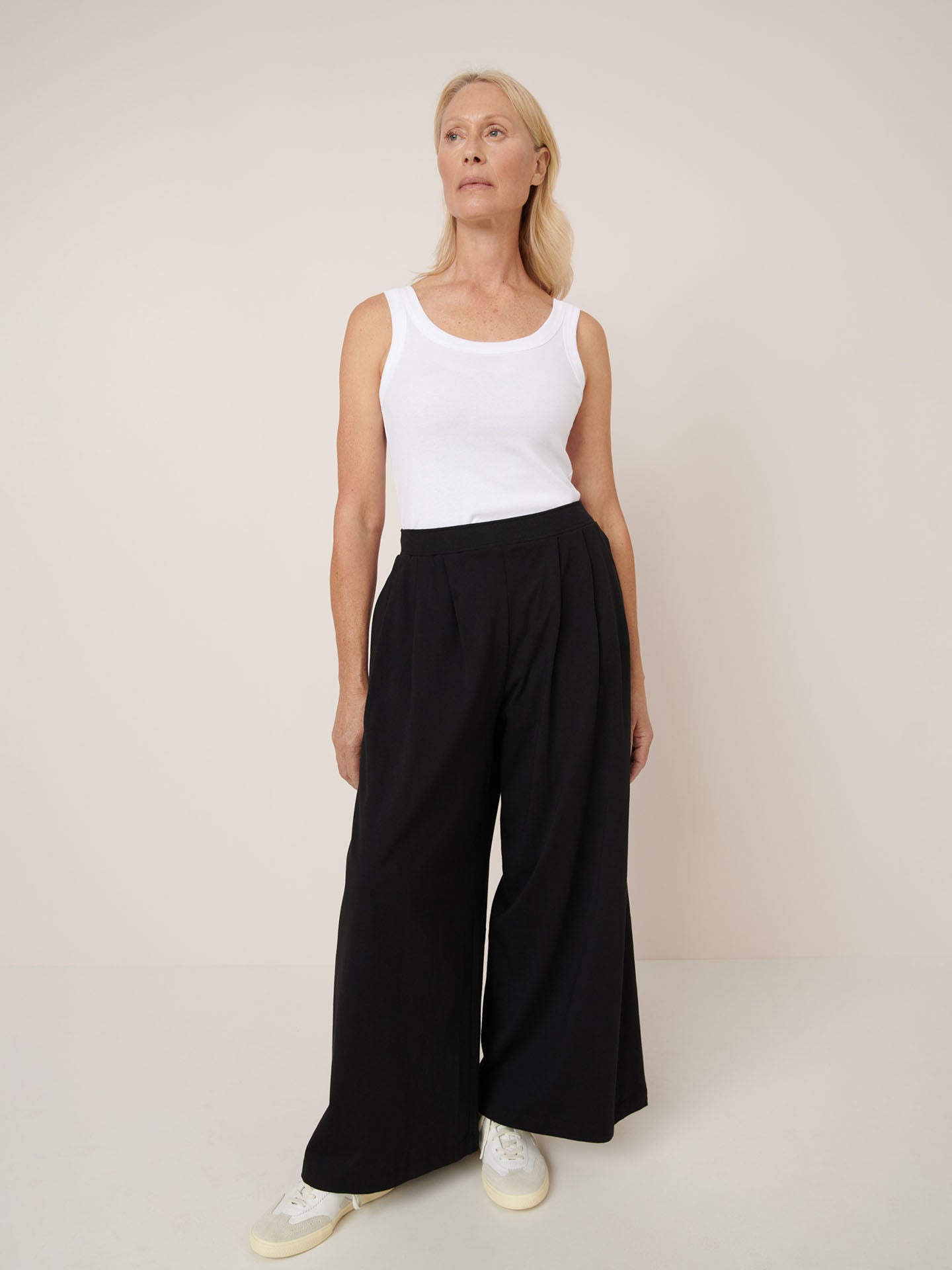 A woman stands against a plain background wearing a white tank top, Kowtow's Drape Pant in black with a wide-leg relaxed fit, and white sneakers.