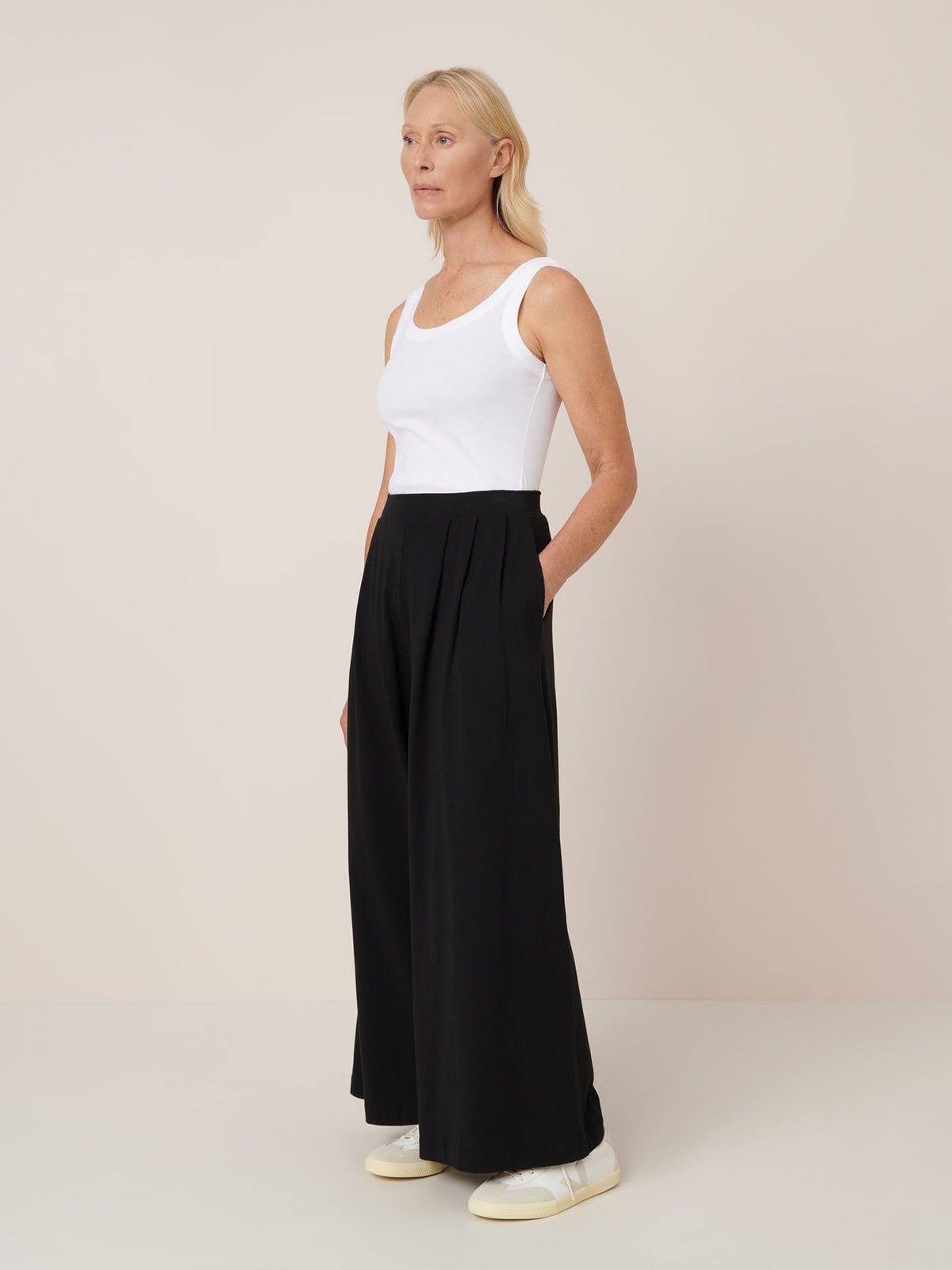 A woman stands against a plain background wearing a white tank top, Kowtow Drape Pant – Black with an elastic waistband, and white sneakers.