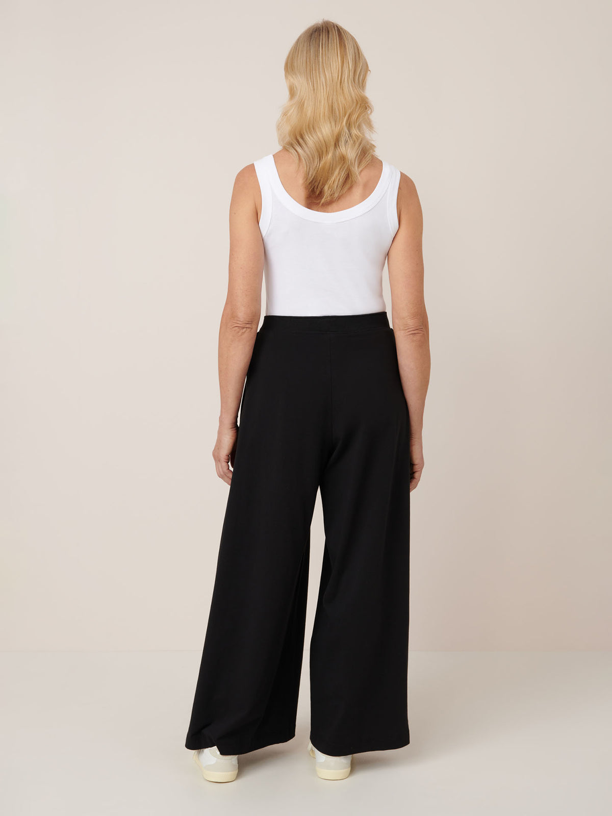A woman with blonde hair, in a white sleeveless top and Kowtow’s Drape Pant – Black, featuring an elastic waistband, stands facing away against a plain light background.