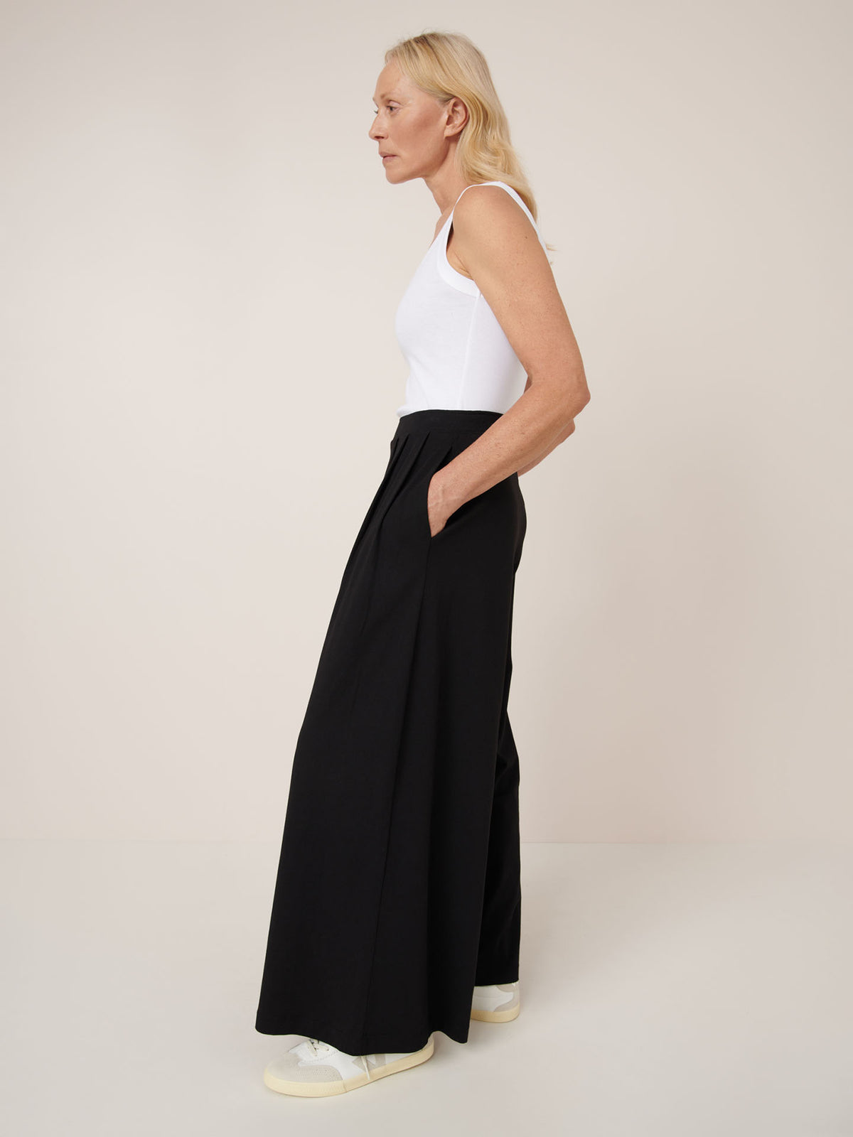 A woman stands in profile against a plain background wearing the Kowtow Drape Pant in black, featuring a high waist and wide legs with an elastic waistband, paired with a white sleeveless top and white sneakers.