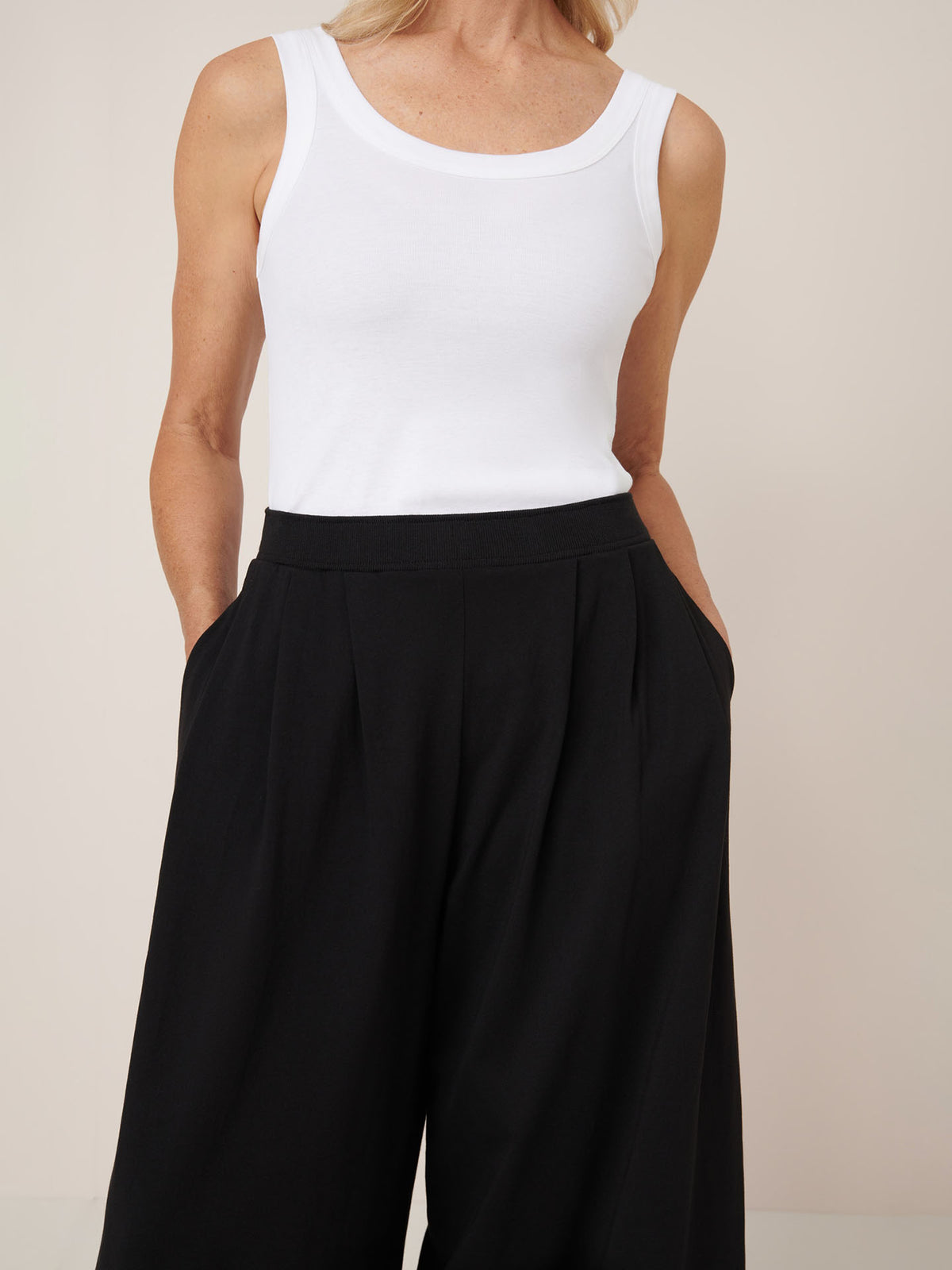 A person wears the Kowtow Drape Pant in black—high-waisted, relaxed fit—with hands in pockets, paired with a sleeveless white top, shown from shoulders to knees.