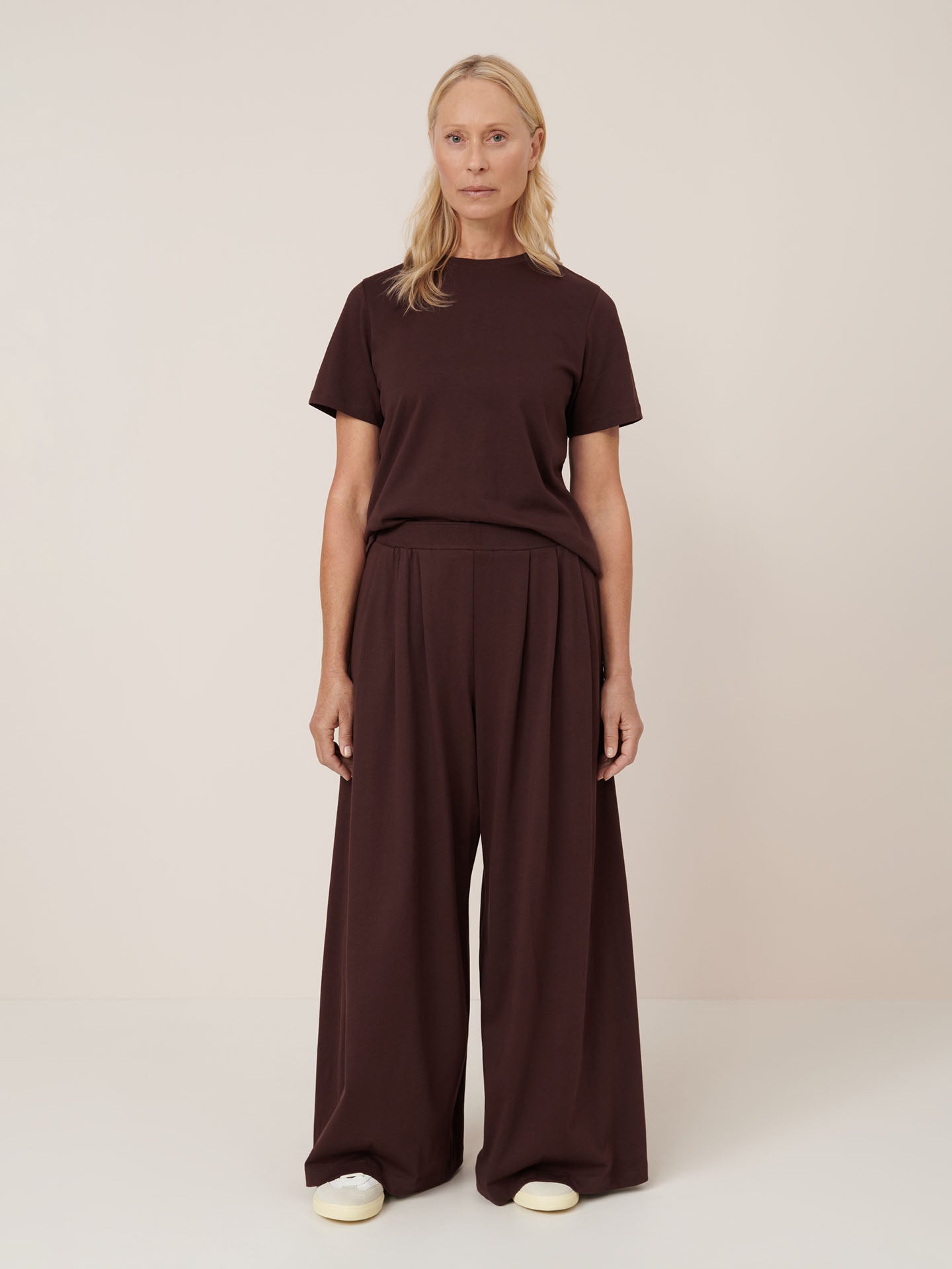 A person stands against a plain background wearing Kowtow’s Drape Pant in Mahogany, styled with a matching dark top and white shoes.