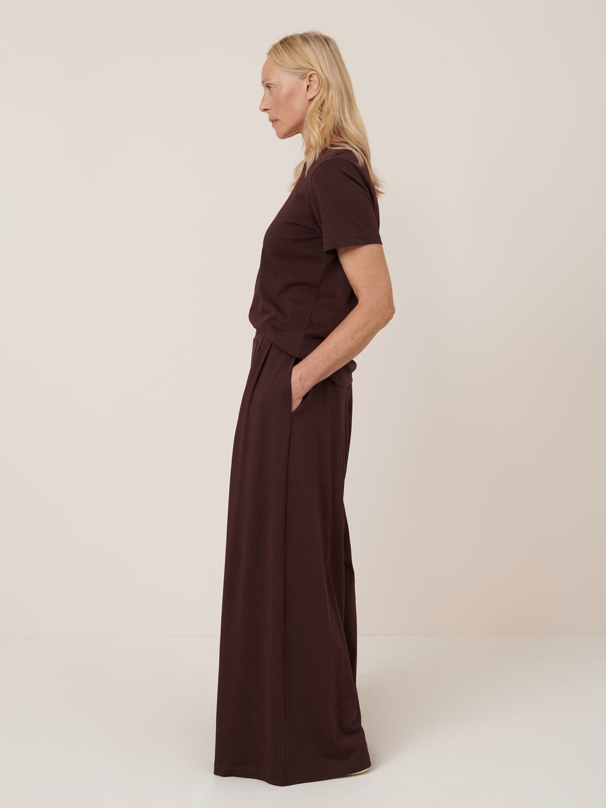 A woman stands in profile against a plain background, wearing the Kowtow Drape Pant in Mahogany, a wide-leg style with an elastic waistband and pockets, paired with a dark brown short-sleeve top.