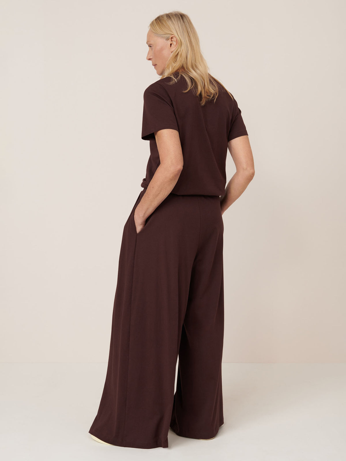 A person with long blond hair, wearing Kowtow’s Drape Pant in Mahogany stands with hands in pockets, facing away from the camera.