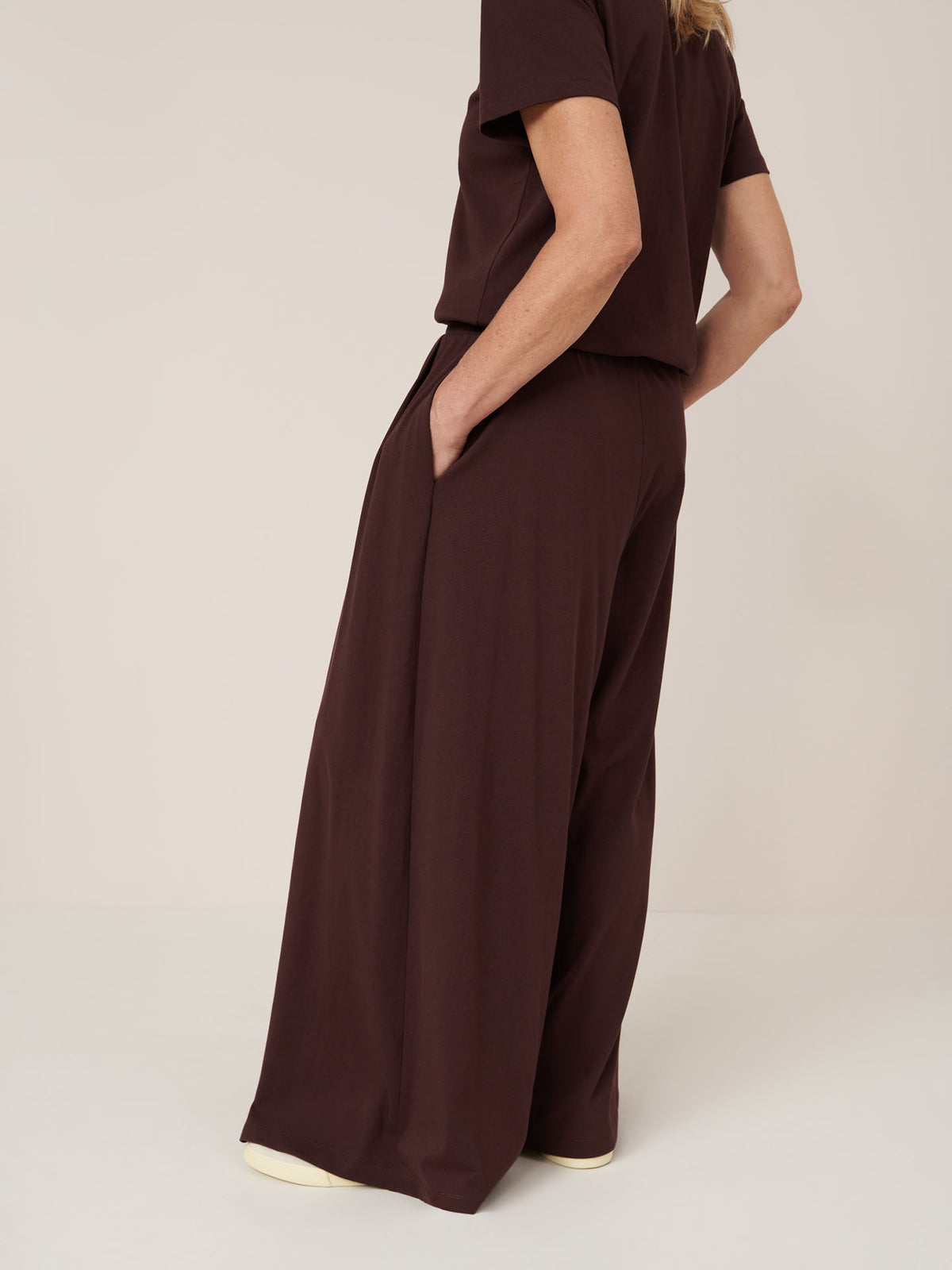 Person wearing Kowtow’s Drape Pant in Mahogany, featuring a wide leg style and elastic waistband, paired with a loose-fitting brown short-sleeve top, standing with one hand in their pocket against a plain background.