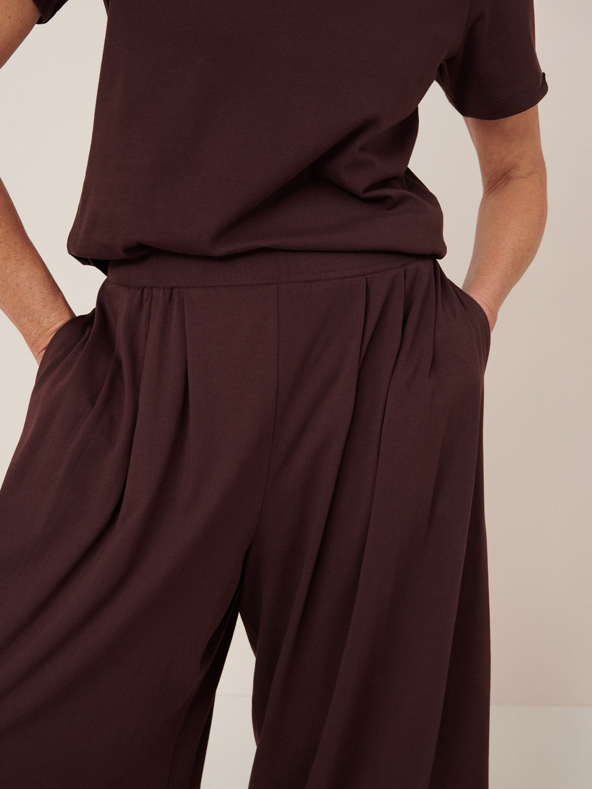Model wears Kowtow&#39;s Drape Pant in Mahogany, a wide-leg elastic waistband pant made from soft knit jersey, paired with a matching dark brown short-sleeve top, hands casually in pockets.