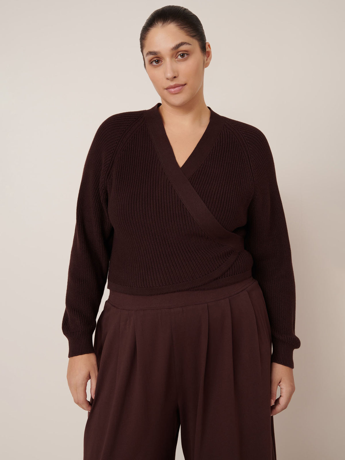 A woman stands against a plain background, wearing Kowtow&#39;s Drape Pant in Mahogany and a dark brown wrap sweater, looking directly at the camera with a neutral expression.