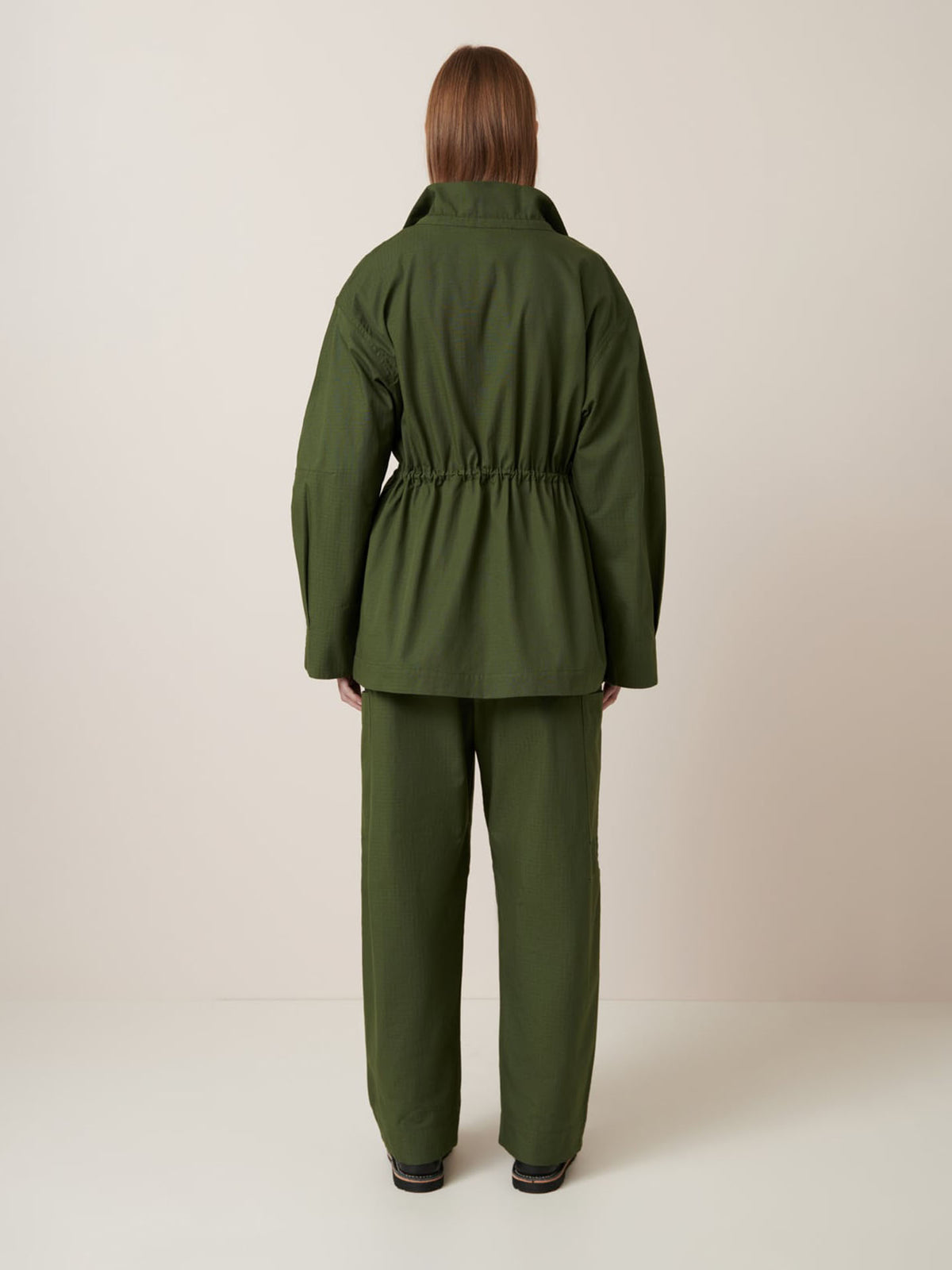 A person stands facing away, wearing the Kowtow Echo Jacket – Forest, a loose-fitting dark green jacket with an elastic waist and matching oversized pants, set against a plain light background.