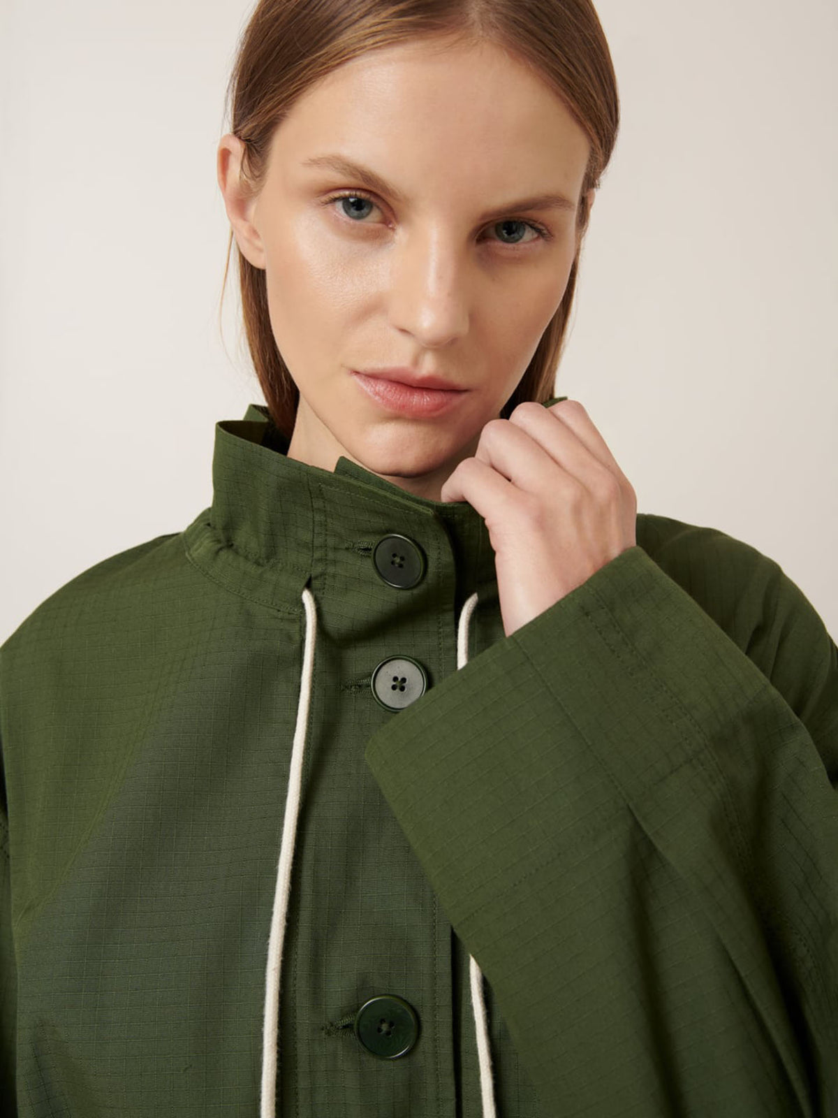 A woman with straight light brown hair wears the Kowtow Echo Jacket – Forest, an oversized green button-up with white drawstrings, and looks directly at the camera.