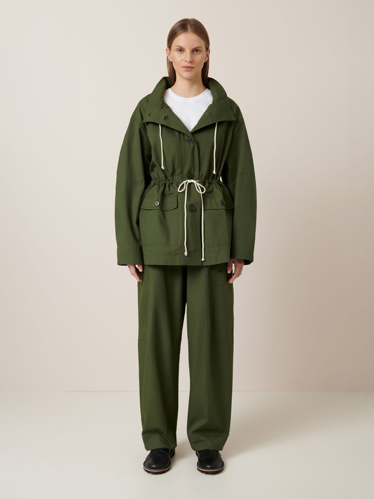 A person stands facing forward against a plain light background, wearing the Kowtow Echo Jacket – Forest with matching loose-fitting pants, black shoes, and a white top.