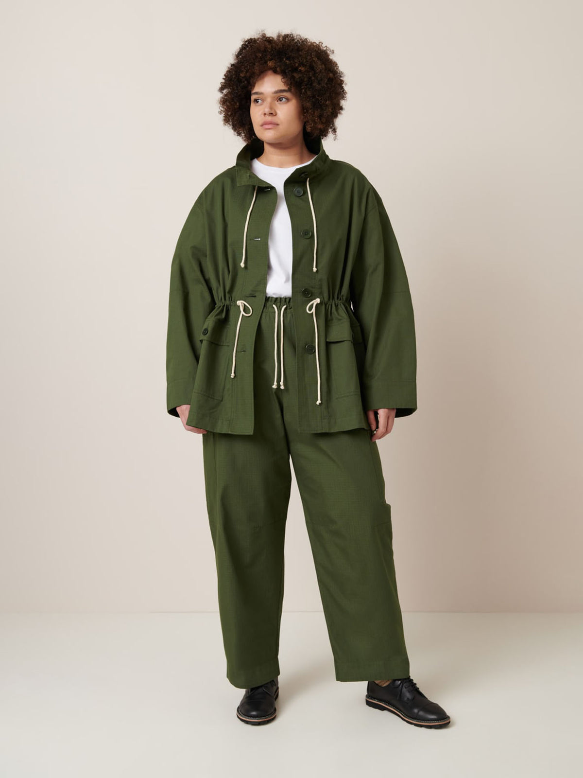 A person stands against a plain background wearing the Kowtow Echo Jacket – Forest in an oversized fit with matching pants, a white t-shirt, and black shoes. Consider product measurements if you prefer not to size down.