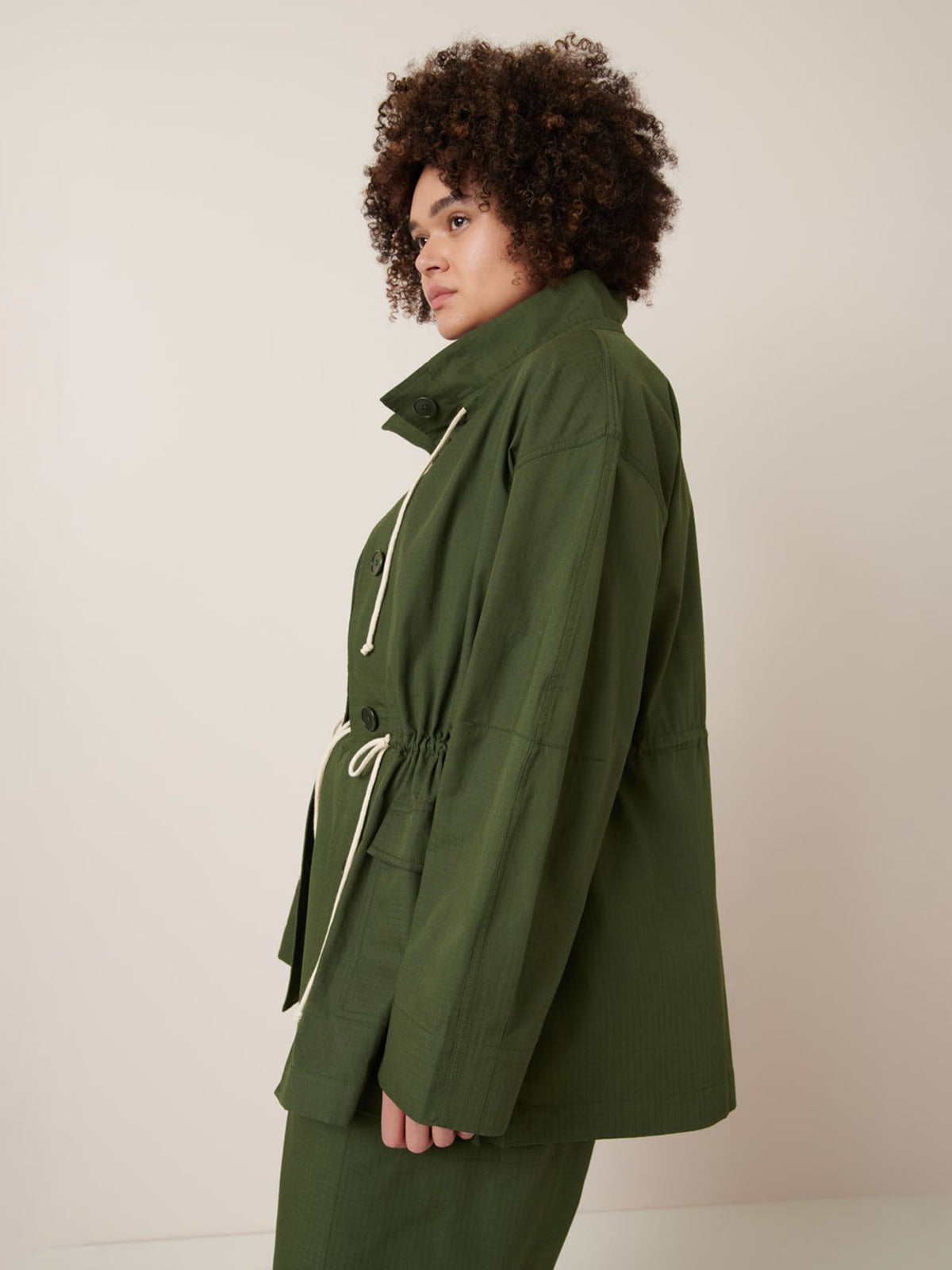 A person with curly hair wears the Echo Jacket – Forest by Kowtow, an olive green button-up with a drawstring waist and oversized fit, standing sideways against a plain light background.