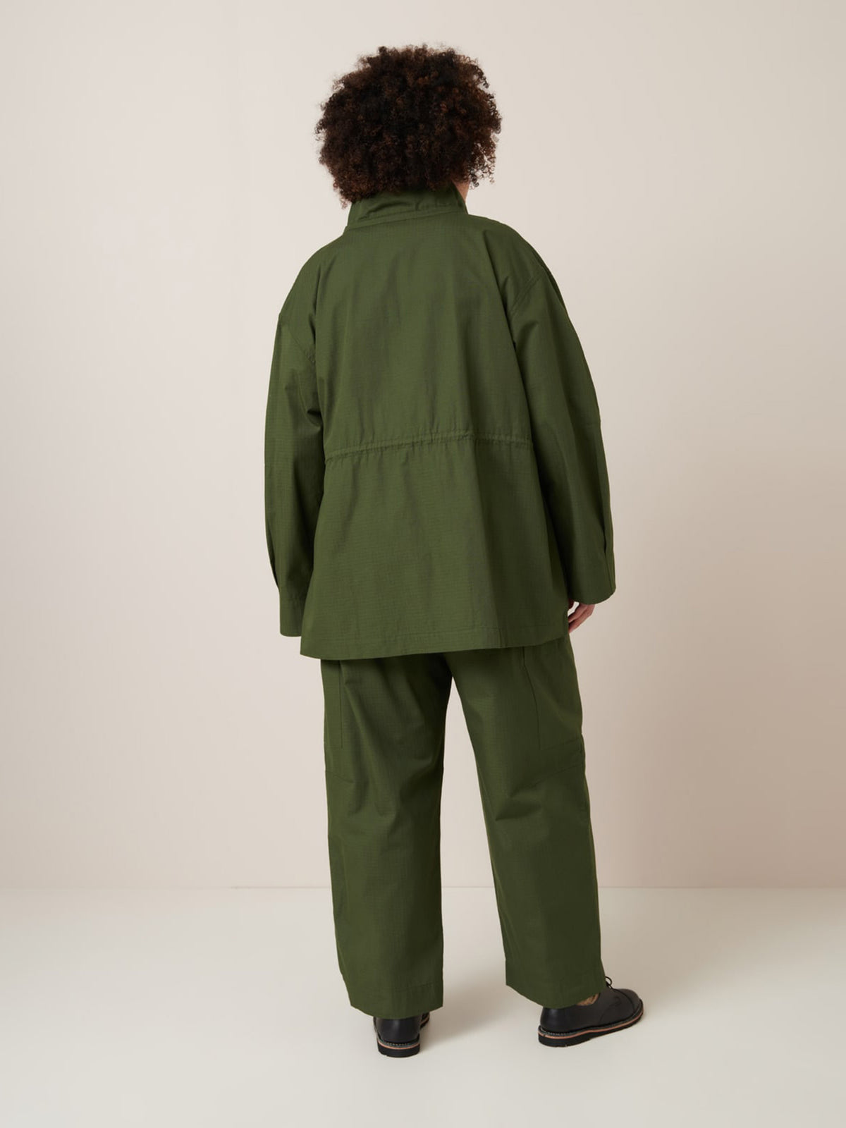 A person stands with their back to the camera, wearing the Kowtow Echo Jacket – Forest in an oversized fit, paired with matching pants and black shoes, set against a plain light background.